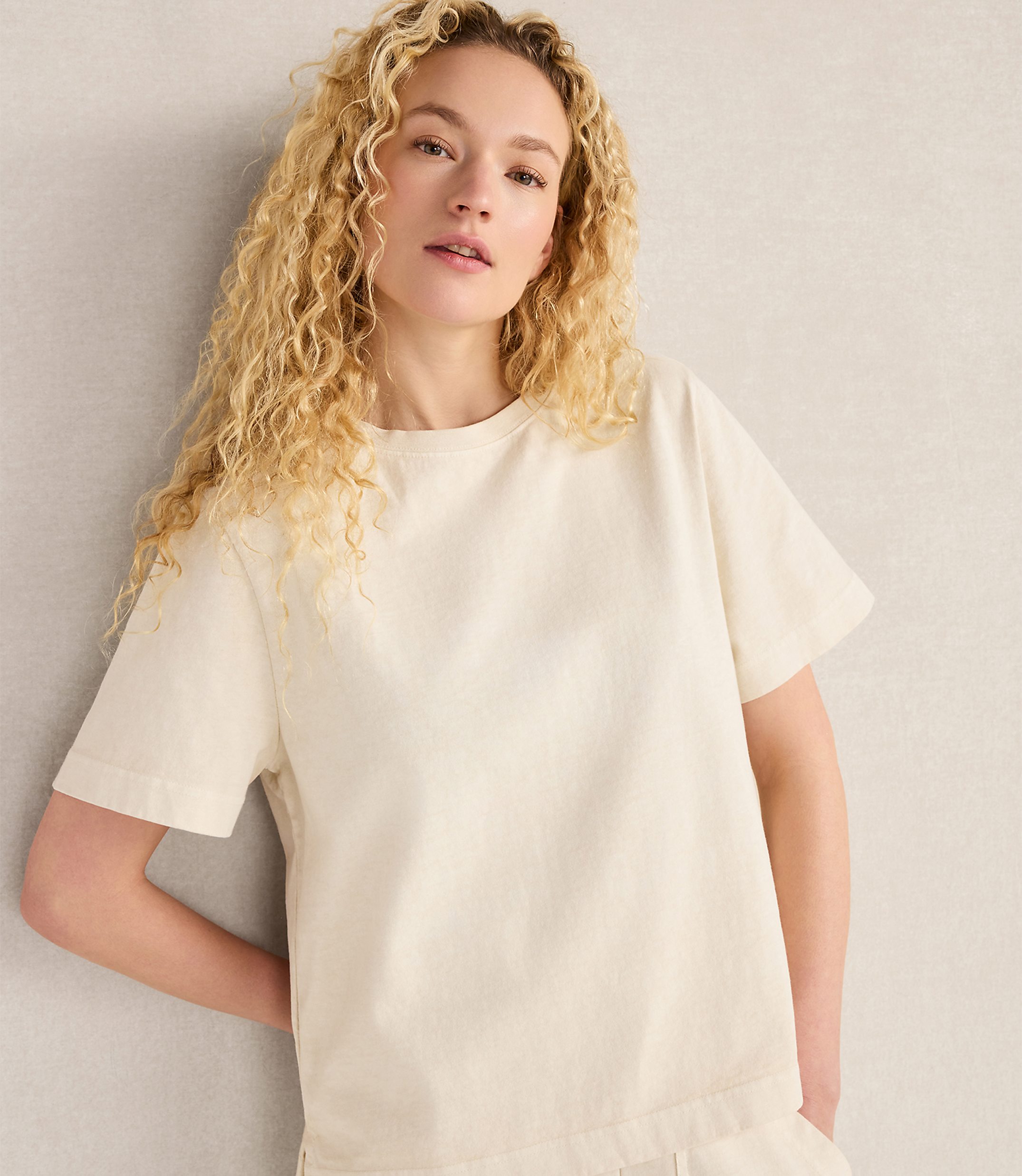 A young woman with curly blonde hair wears a cream-colored t-shirt, leaning against a neutral-toned wall.