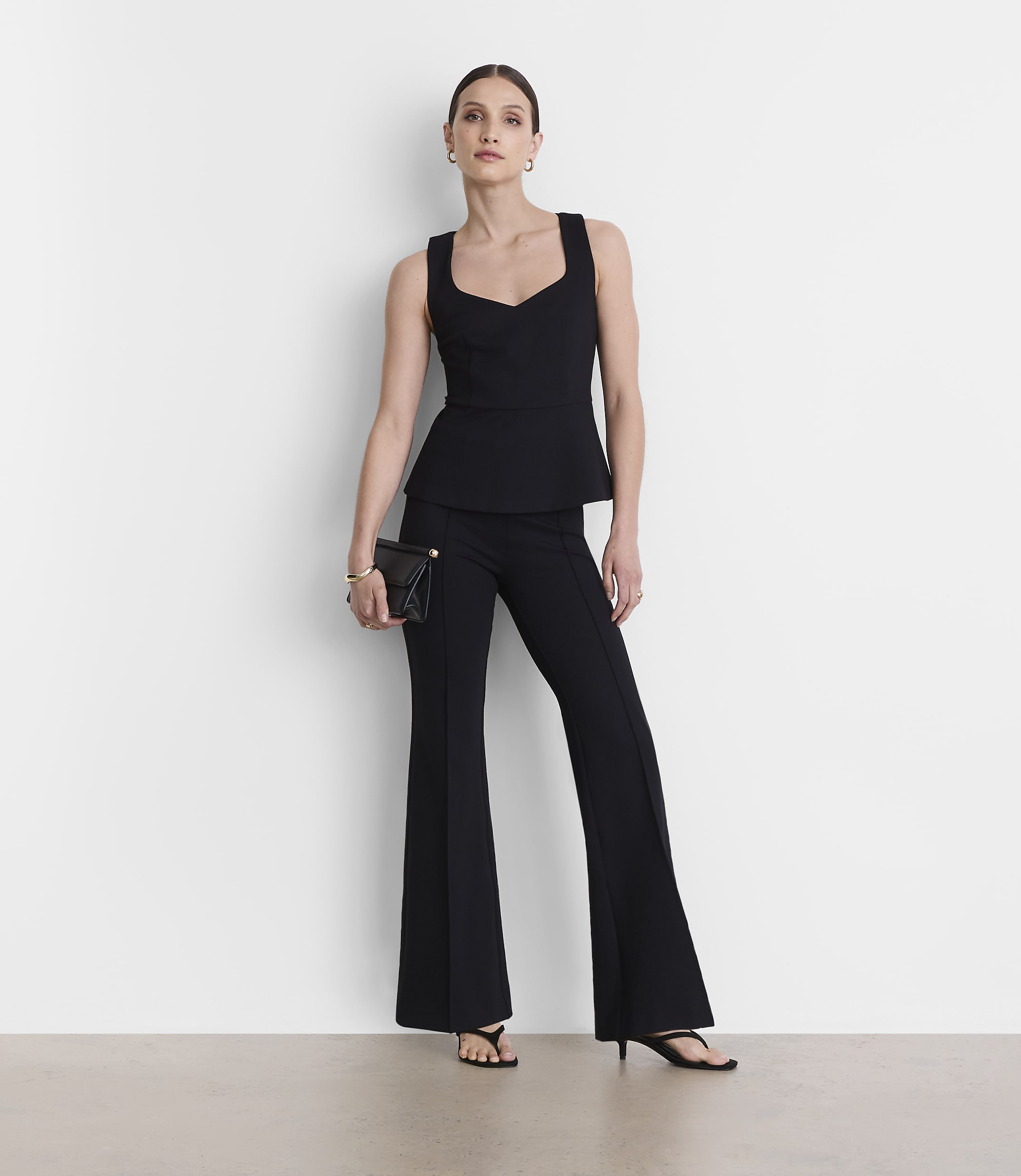 A woman in a black peplum top and flared pants stands against a plain white background, holding a black clutch.