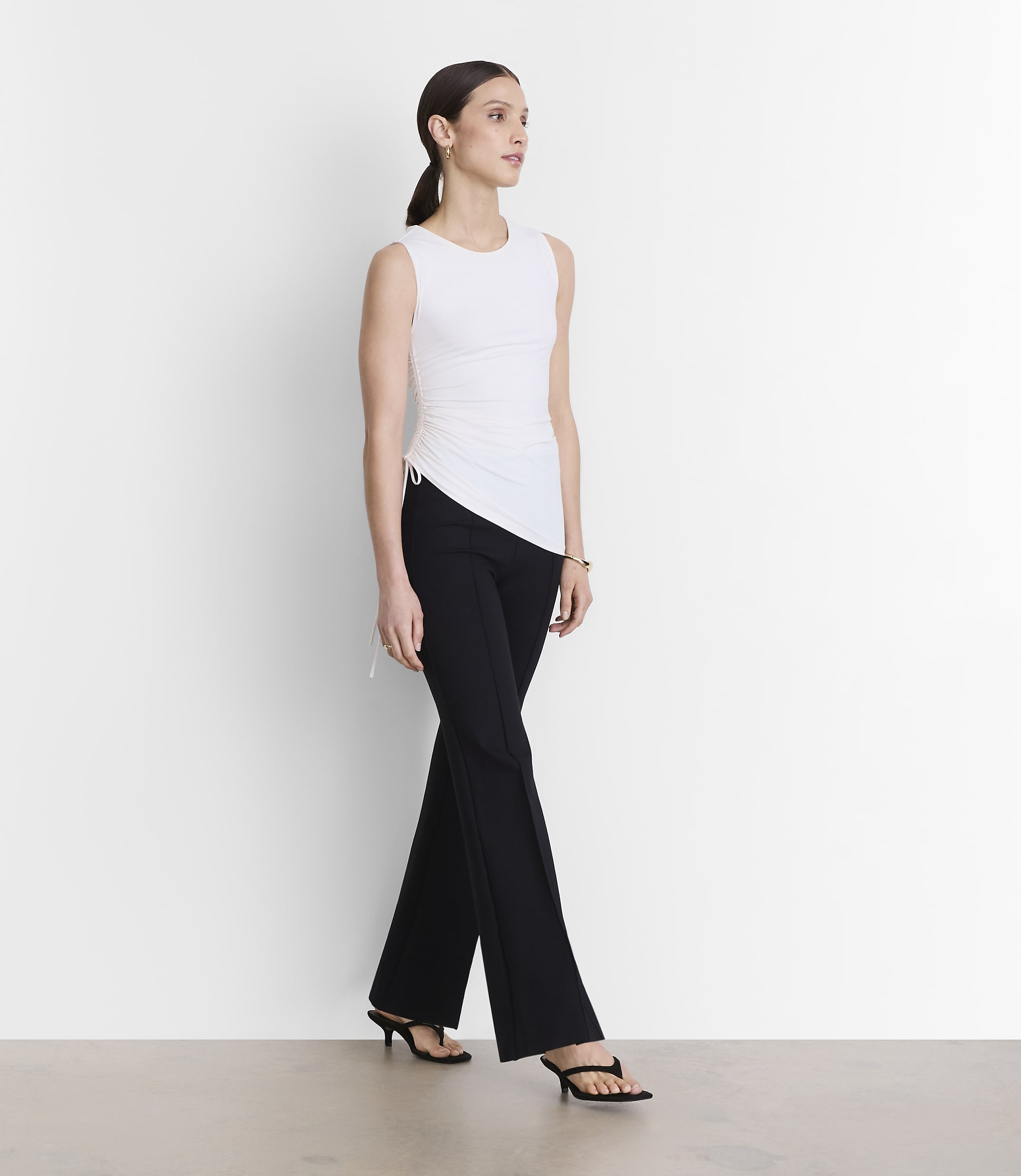 A woman in a white sleeveless top and black pants stands against a plain white background.