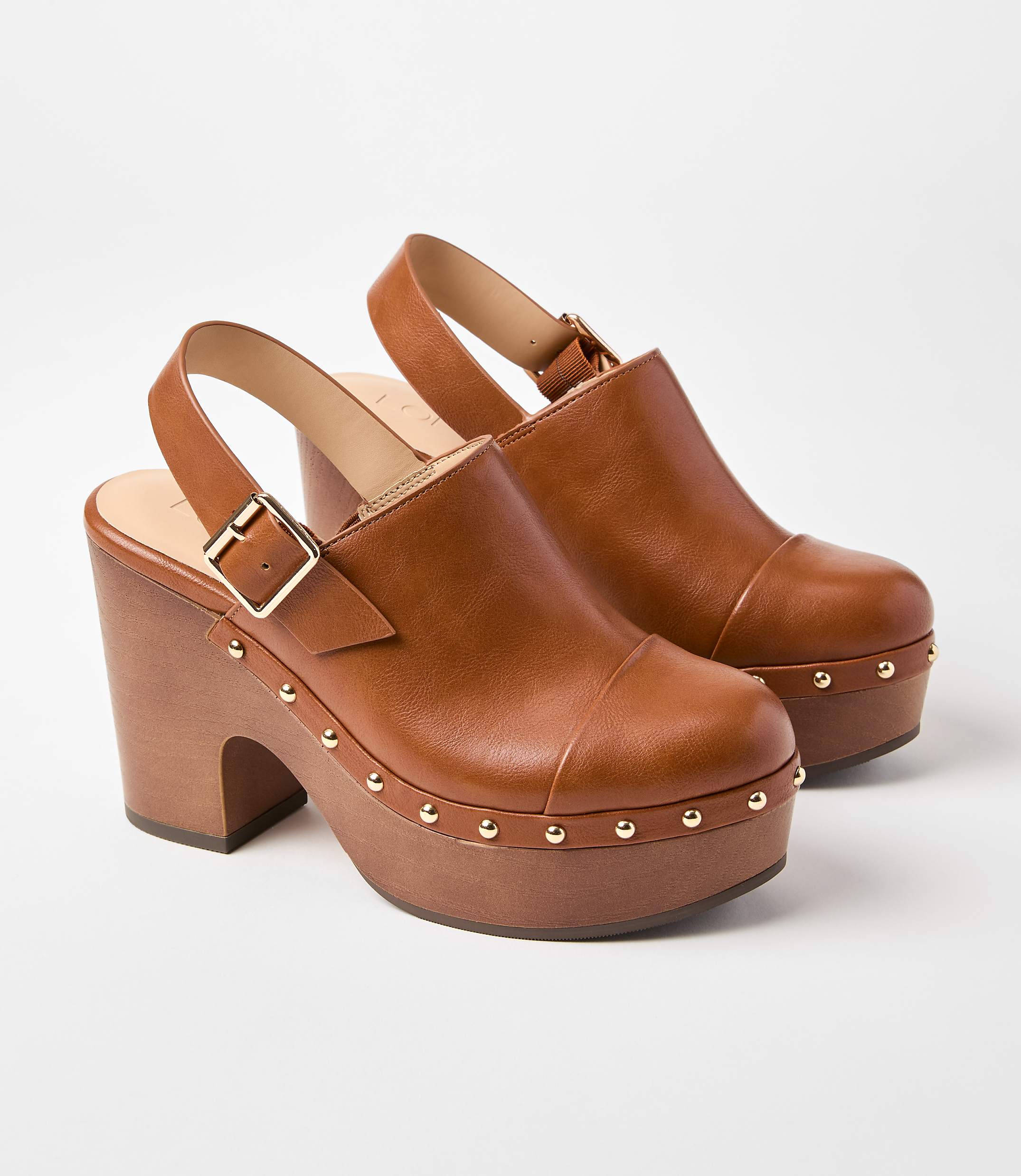 A pair of brown leather clogs with wooden platform soles and gold stud detailing are displayed against a plain white background.