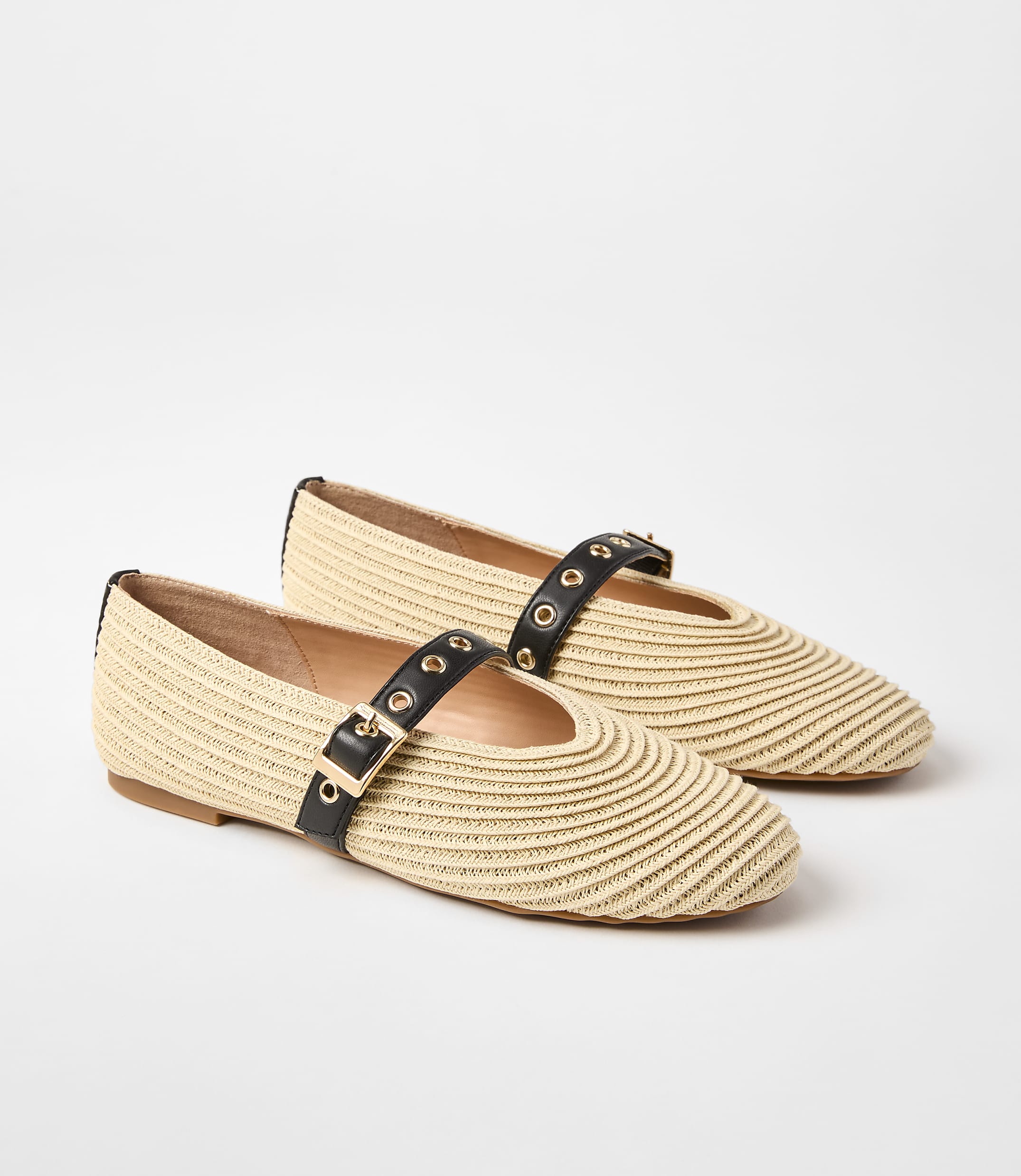 A pair of woven straw ballet flats with black straps and gold buckles rests on a plain white background.