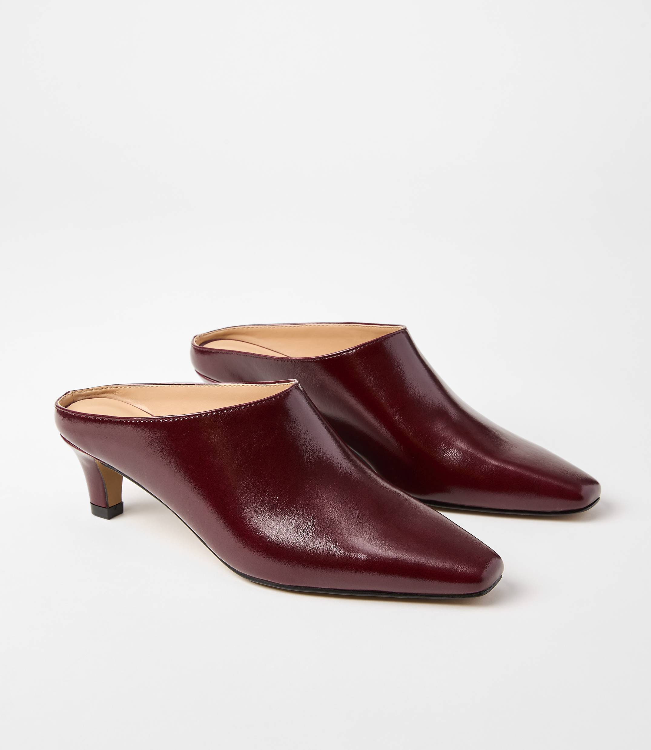 A pair of burgundy leather mules with pointed toes and low heels rests on a plain white background.