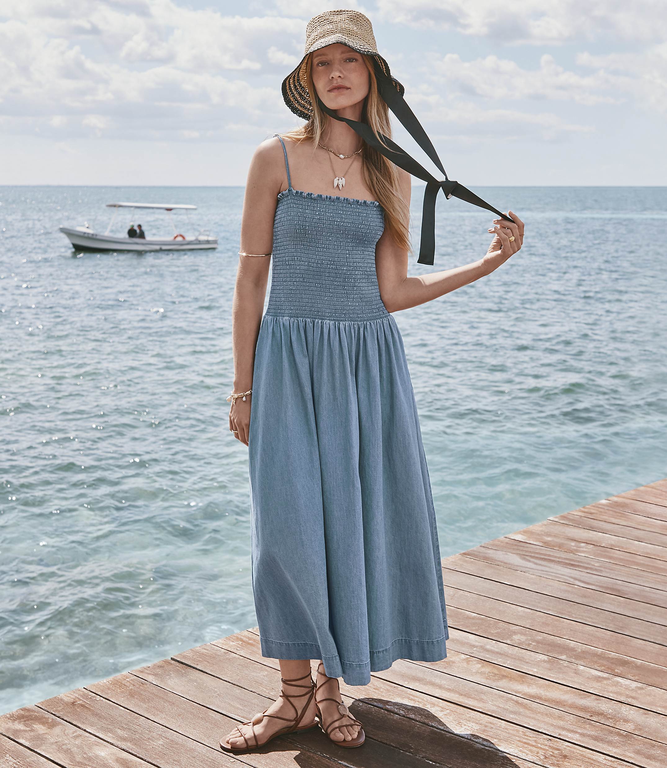 A woman in a blue dress and straw hat stands on a wooden pier with a boat in the calm ocean behind her.
