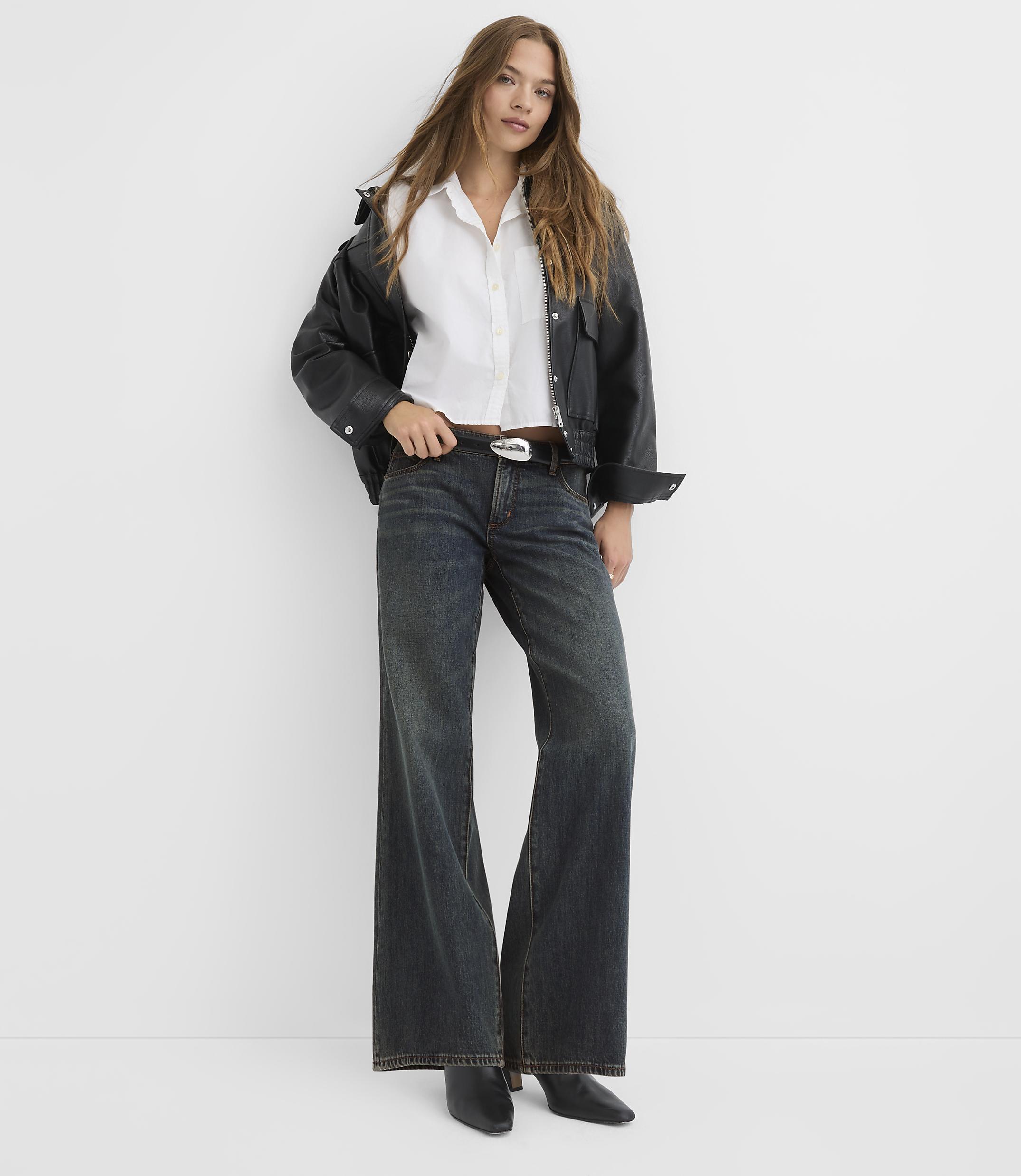 A woman in a white shirt, black jacket, and wide-leg jeans stands against a plain white background.