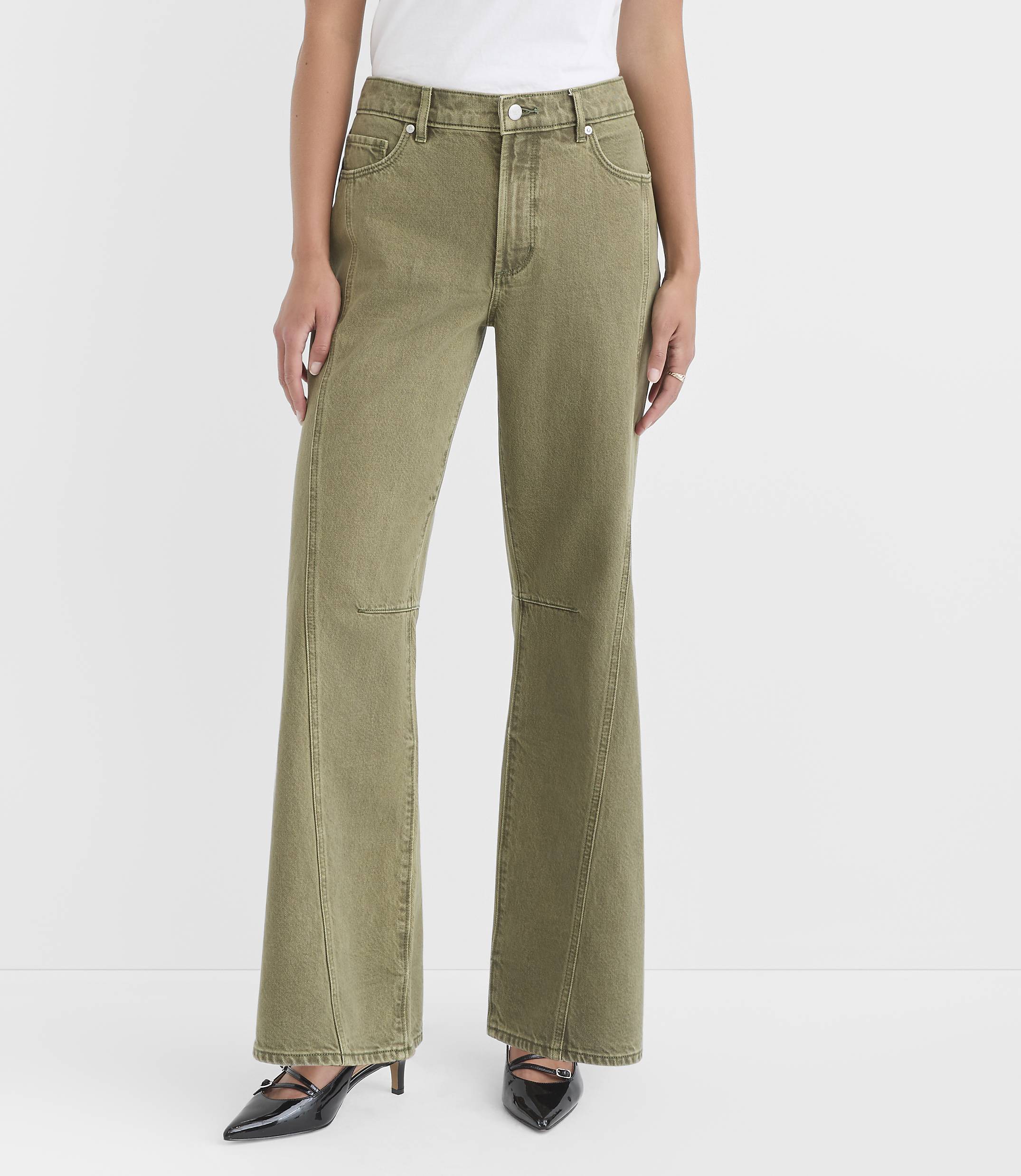 A woman in olive green flared jeans and black heels stands against a plain white background.