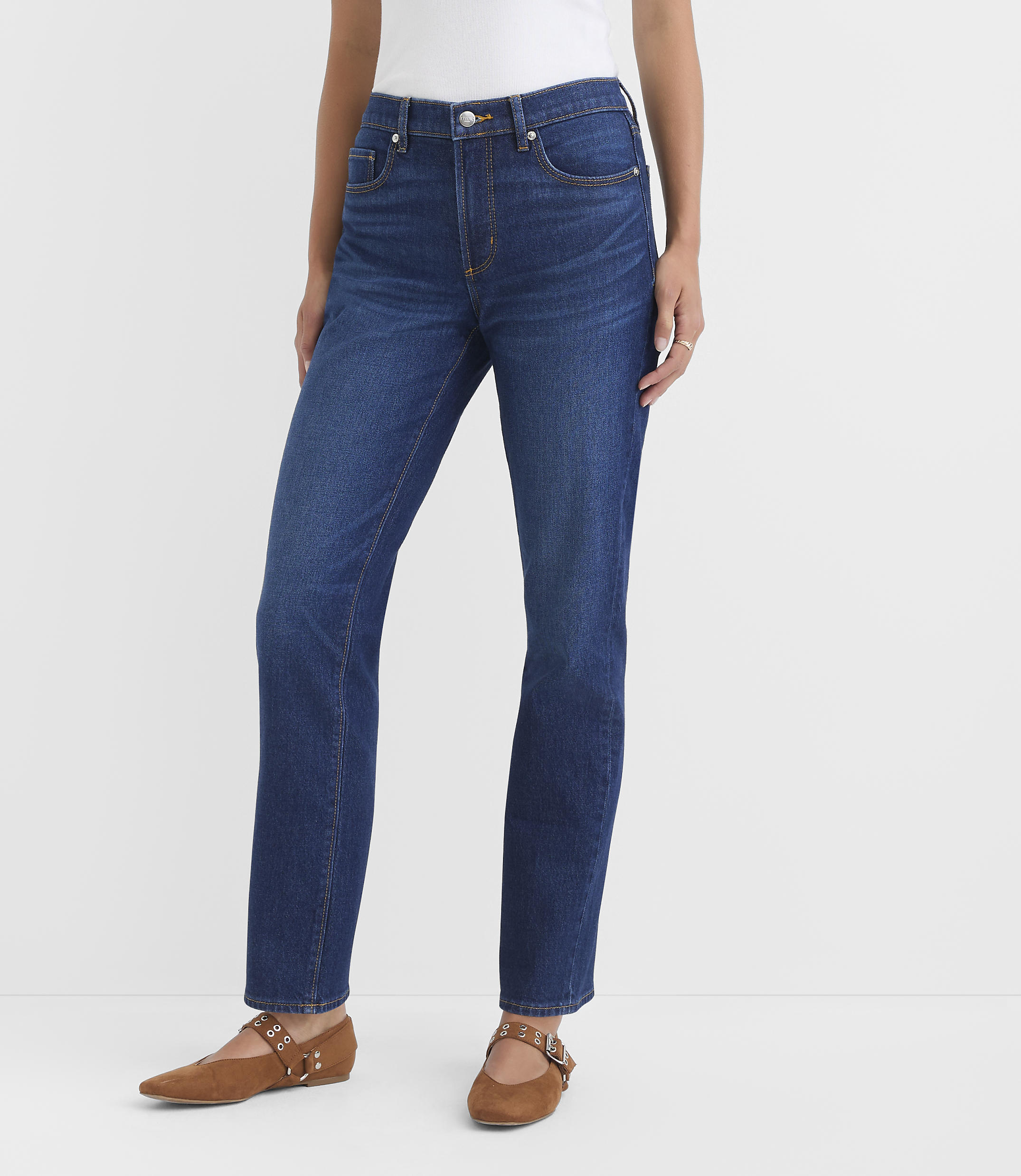 A woman in dark wash jeans and brown flats stands against a plain white background.