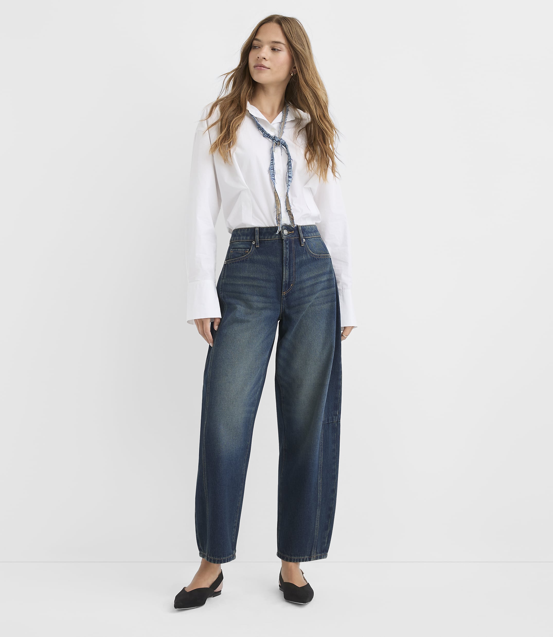 A woman in a white shirt and wide-leg jeans stands against a plain white background.