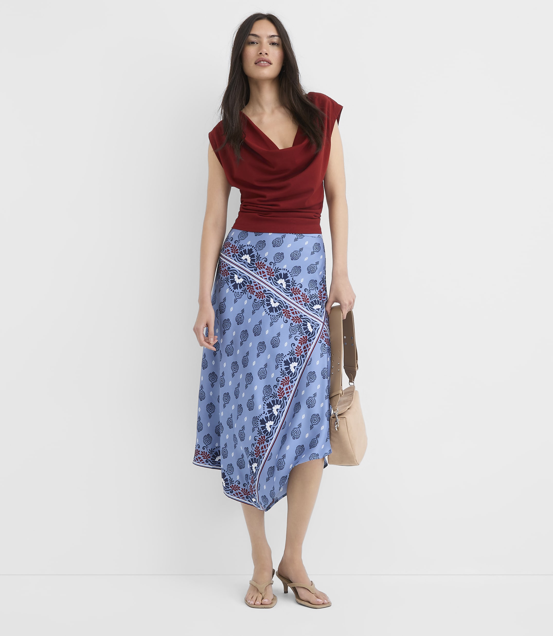 A woman in a rust-colored cowl-neck top and a blue patterned asymmetrical skirt stands against a plain white background, holding a beige crossbody bag.