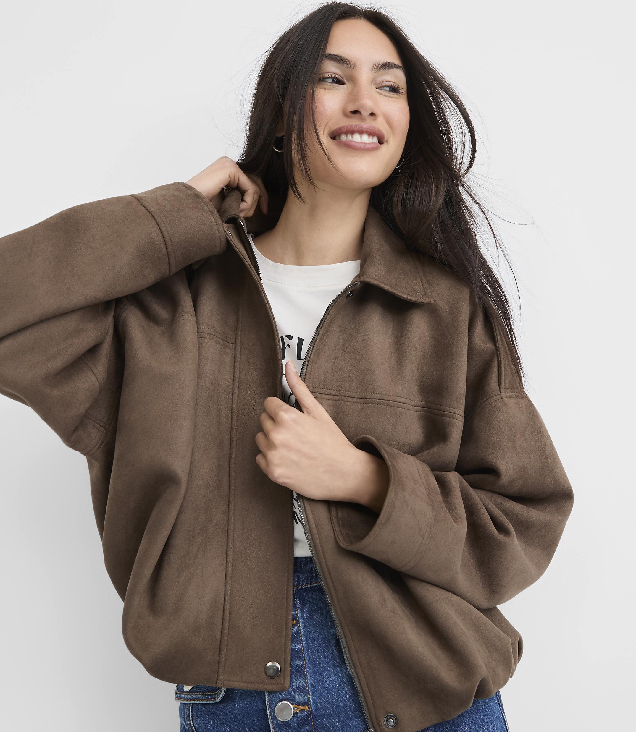 A smiling woman in a brown jacket and jeans stands against a plain white background.