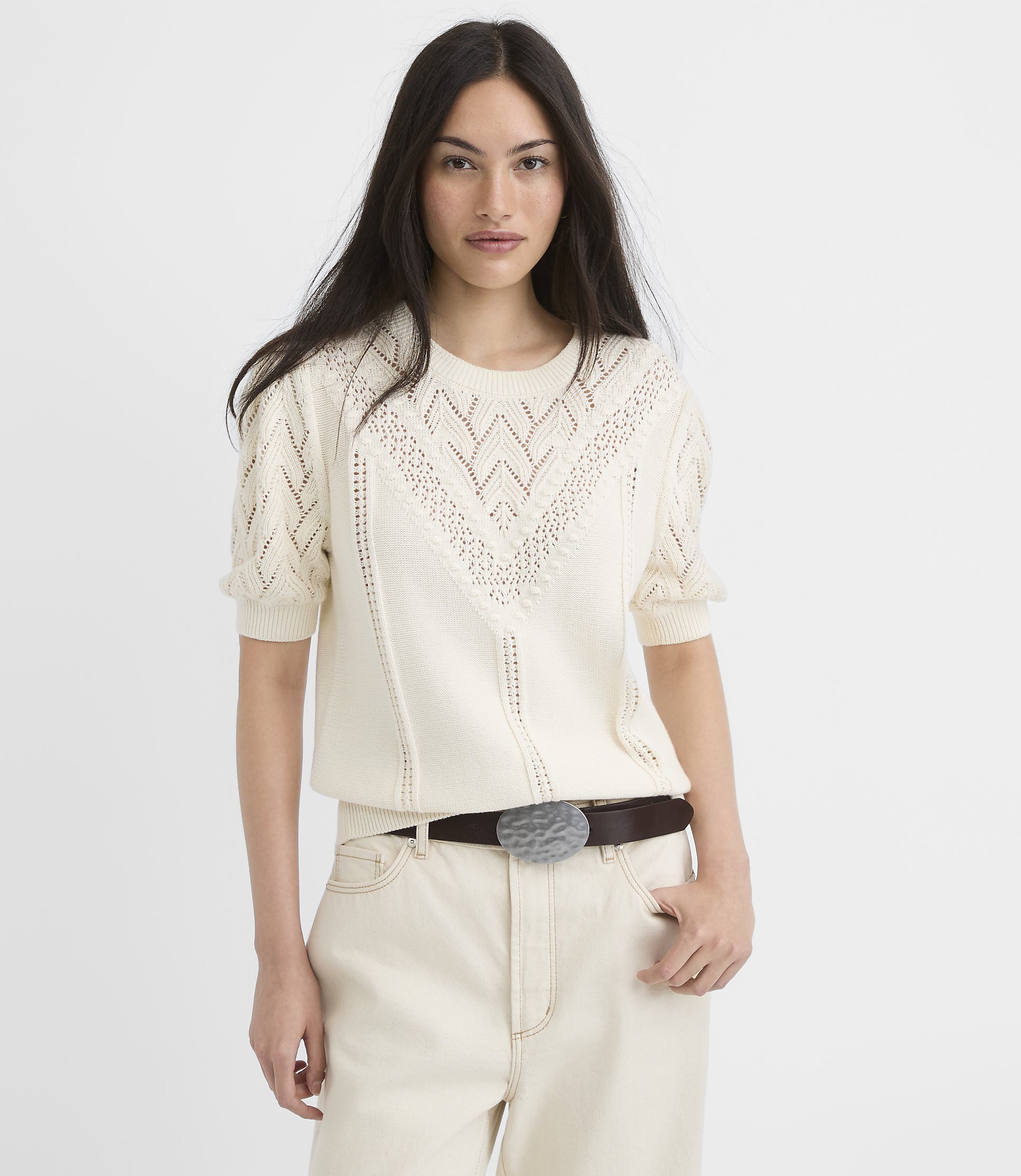 A woman in a cream-colored knit sweater and light-colored pants stands against a plain white background.