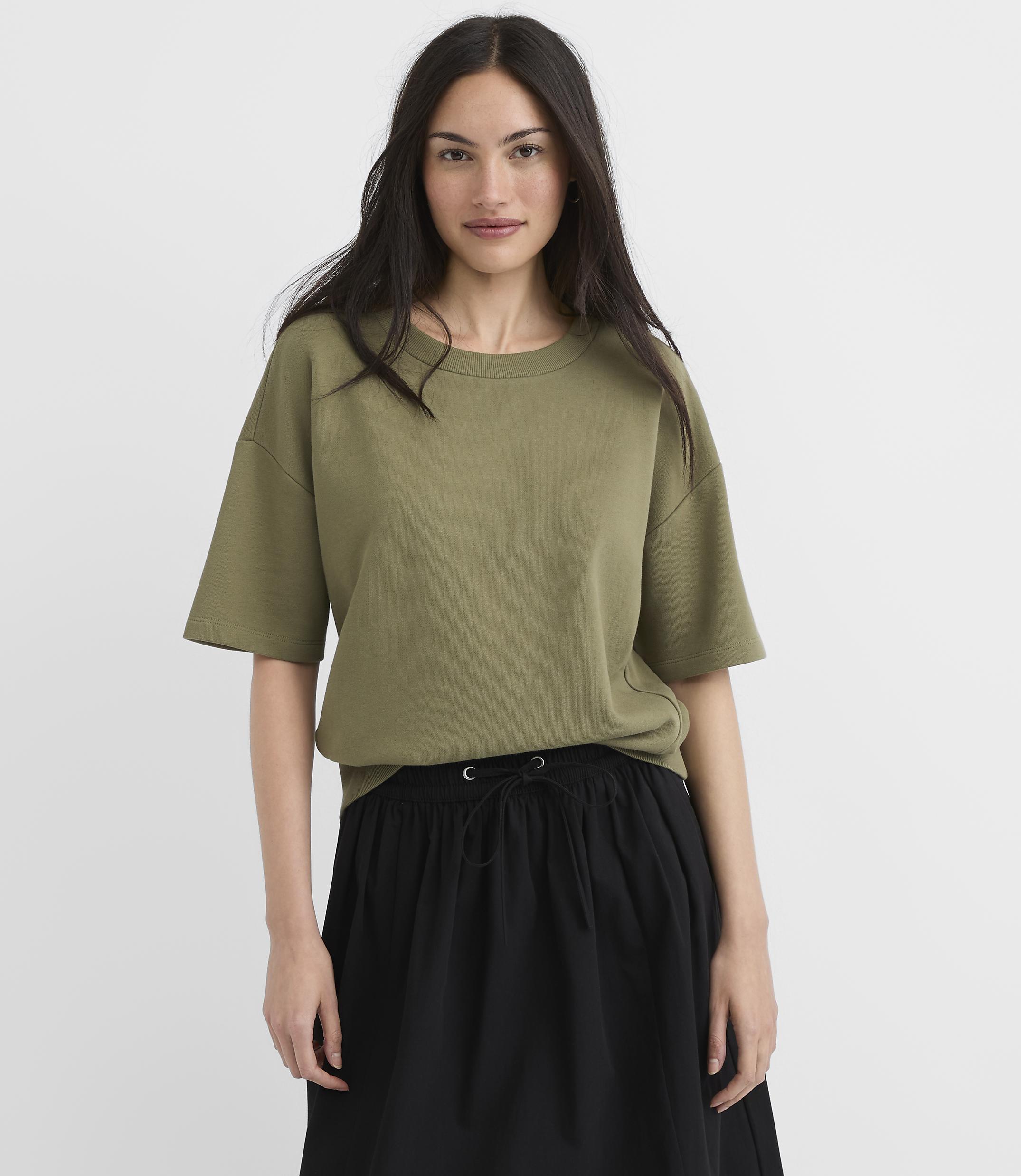 A woman in a green t-shirt and black skirt stands against a plain white background.