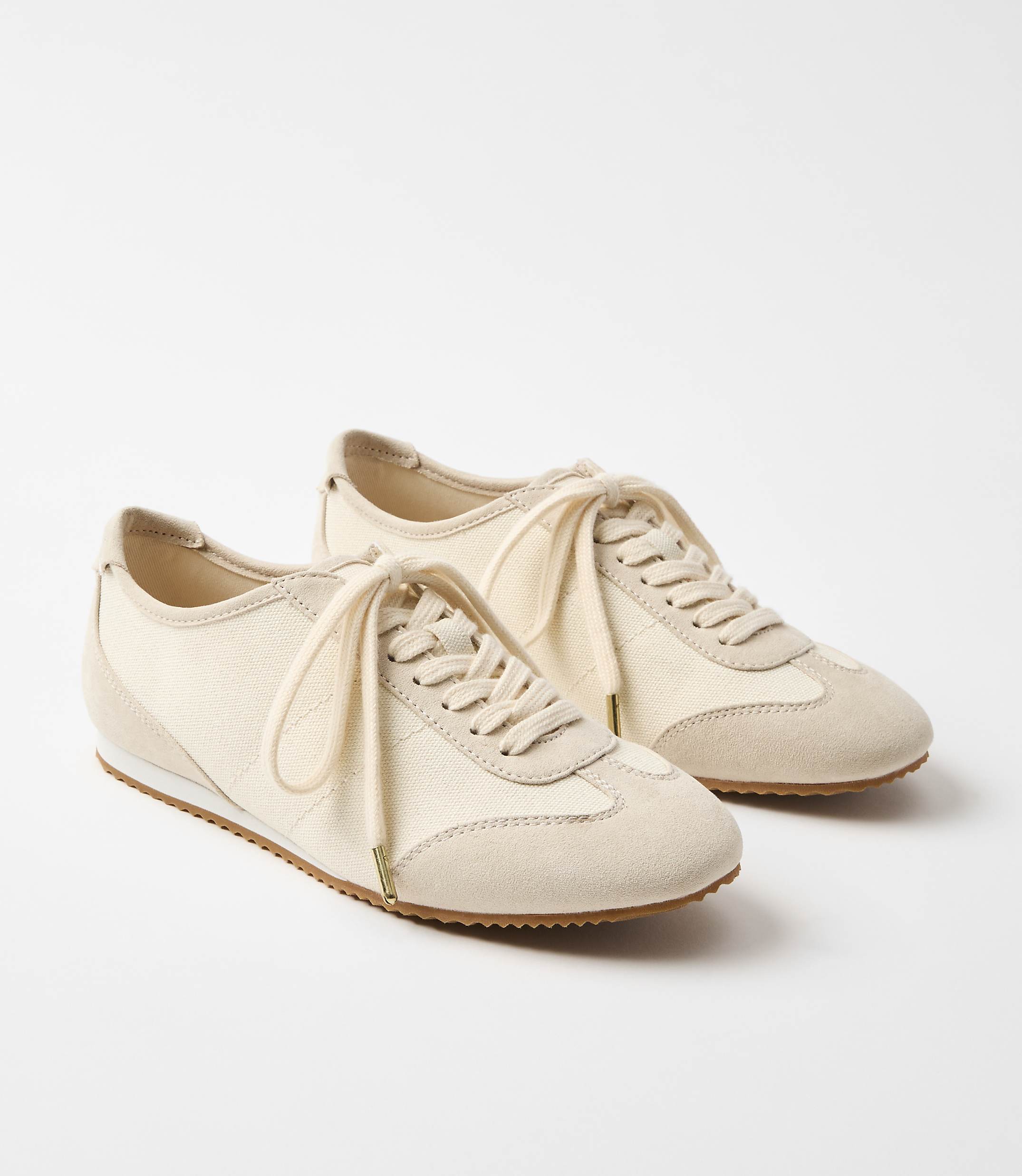 A pair of cream-colored canvas and suede sneakers with tan laces rests on a white surface against a plain white background.