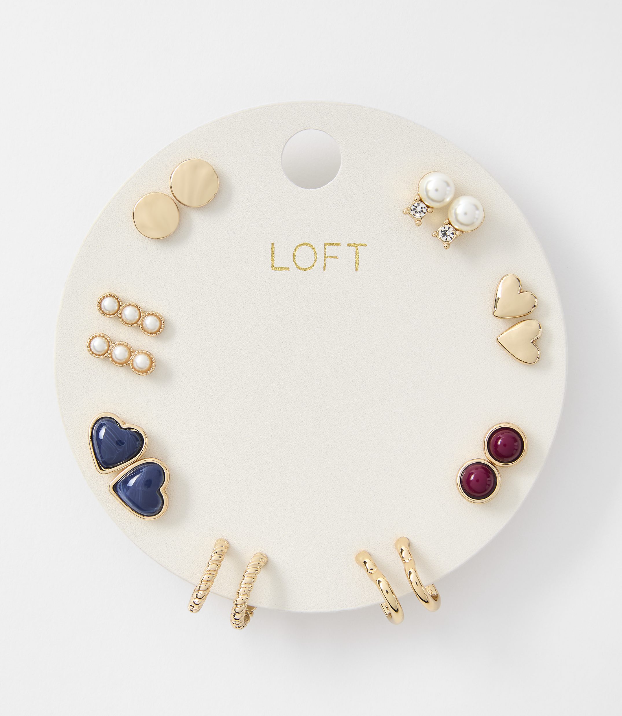 A white circular card displays a collection of gold and pearl earrings, including studs, hoops, and heart shapes, against a plain white background.