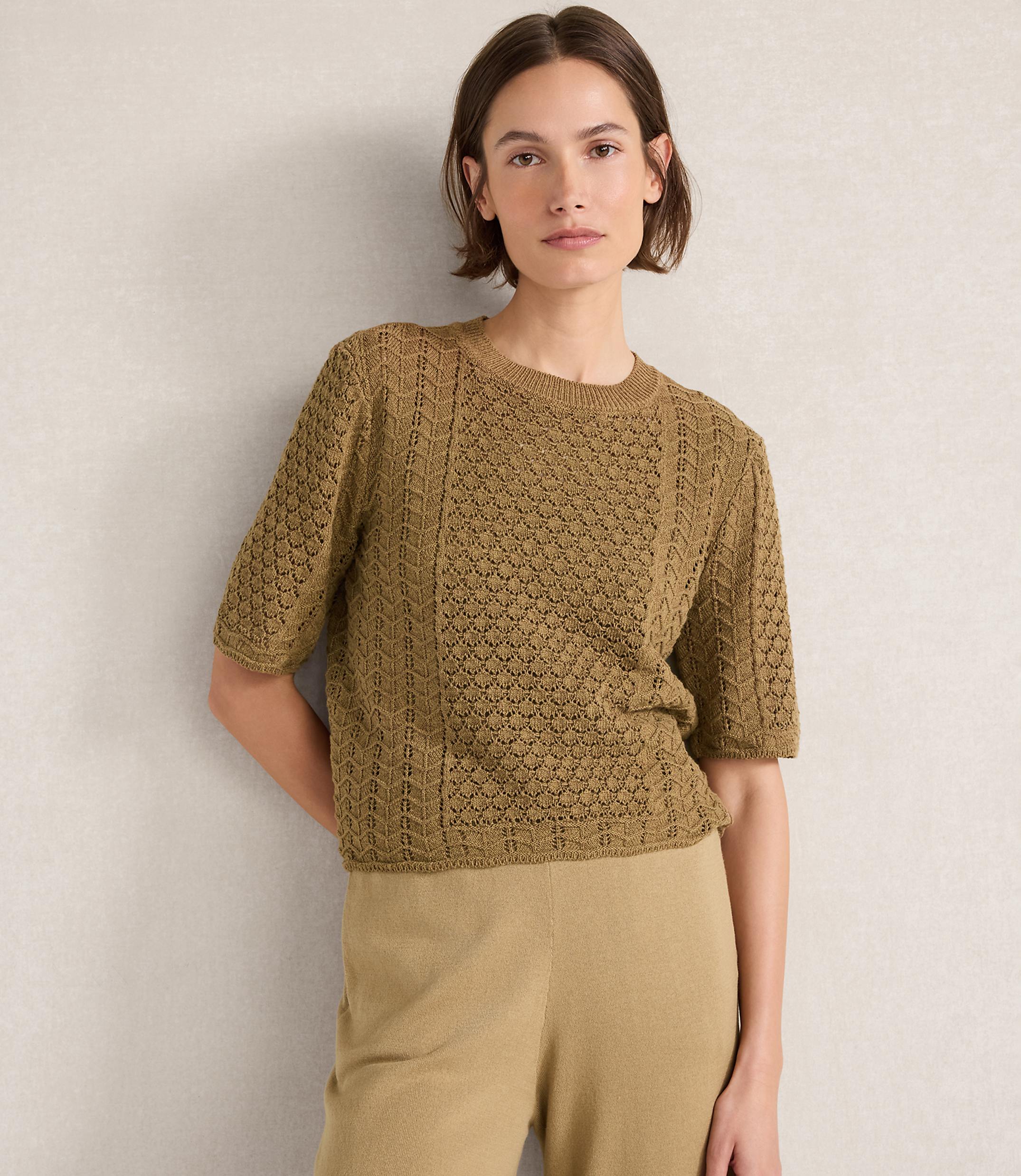 A woman in a textured olive green sweater and tan pants stands against a neutral, textured wall.