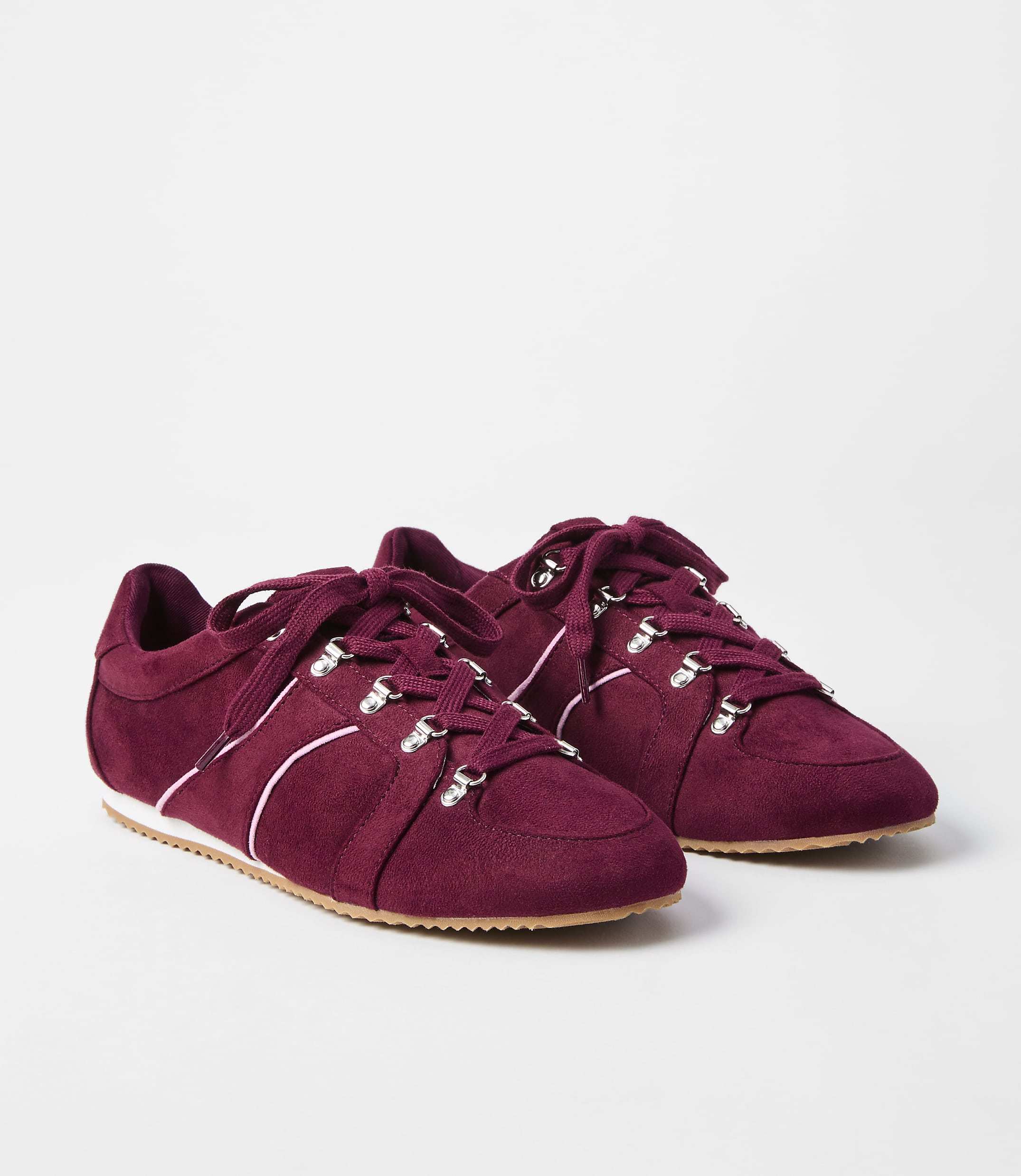 A pair of burgundy suede sneakers with white laces and silver eyelets rests on a white surface against a plain white background.