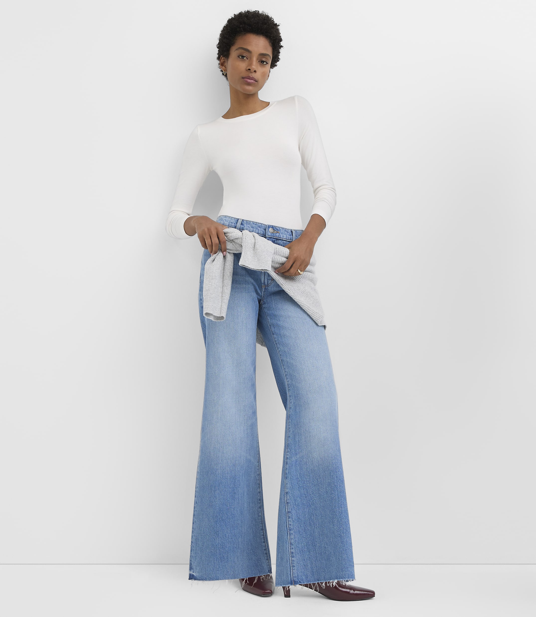 A woman in a white long-sleeved shirt and wide-leg jeans stands against a plain white background.