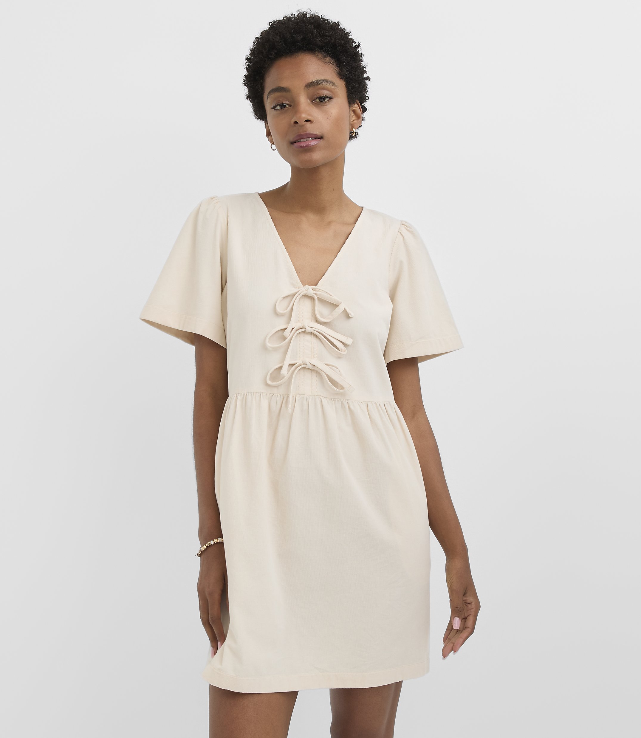A woman in a cream-colored dress with flutter sleeves and a tie-front neckline stands against a plain white background.