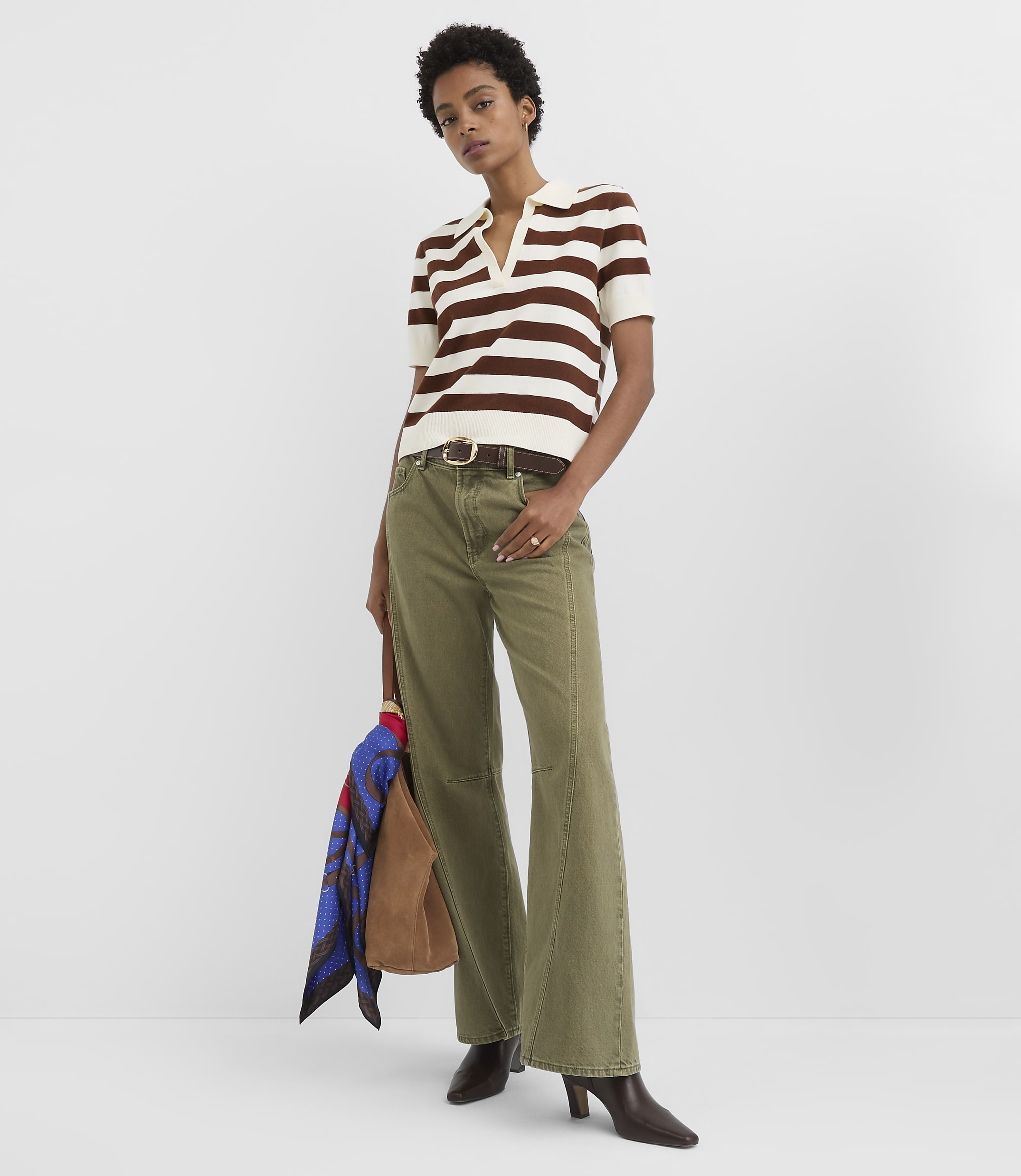 A woman in a striped polo shirt and olive green flared pants stands against a plain white background, holding a brown bag with a colorful scarf.