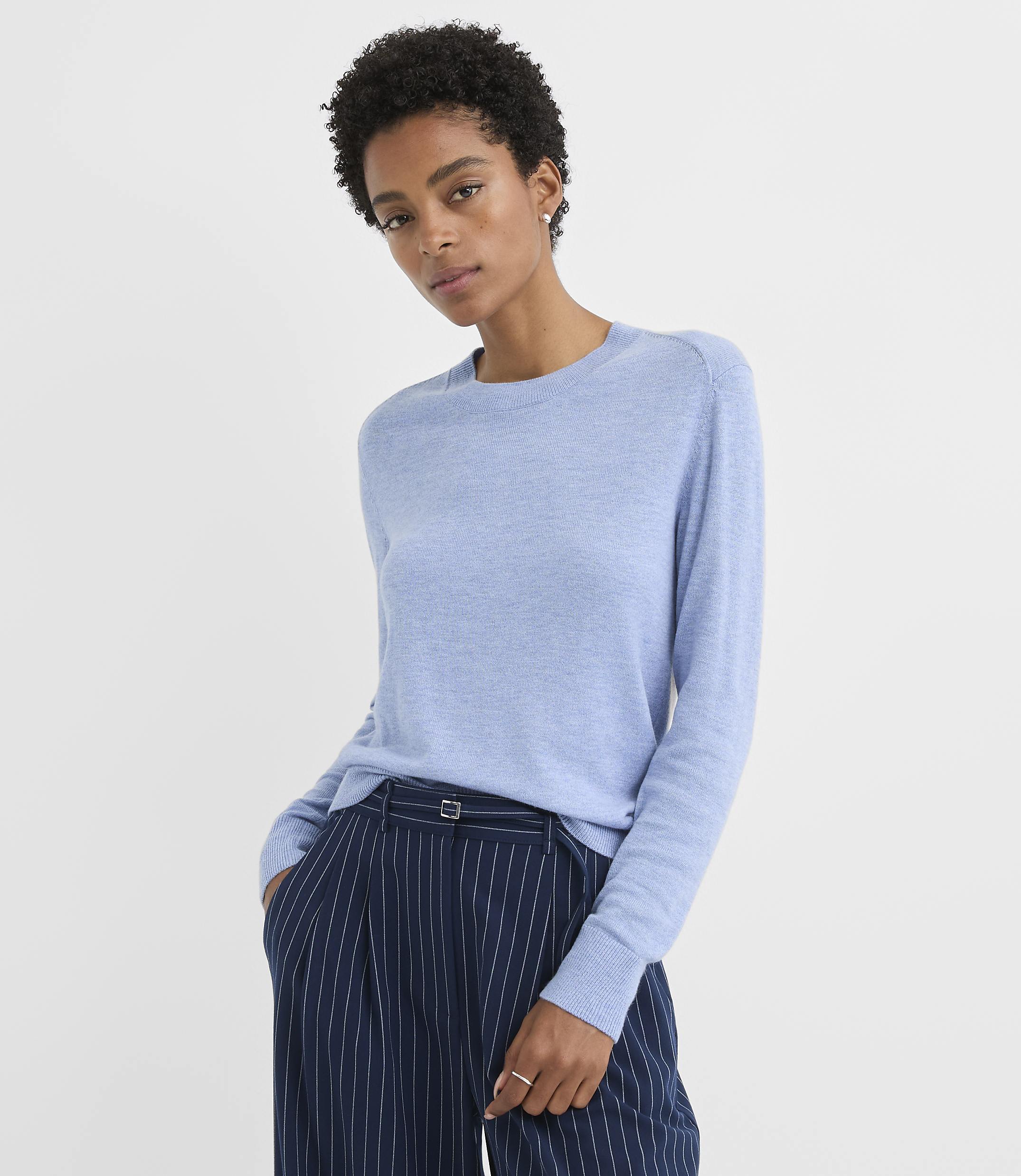 A woman in a light blue sweater and pinstripe pants stands against a plain white background.