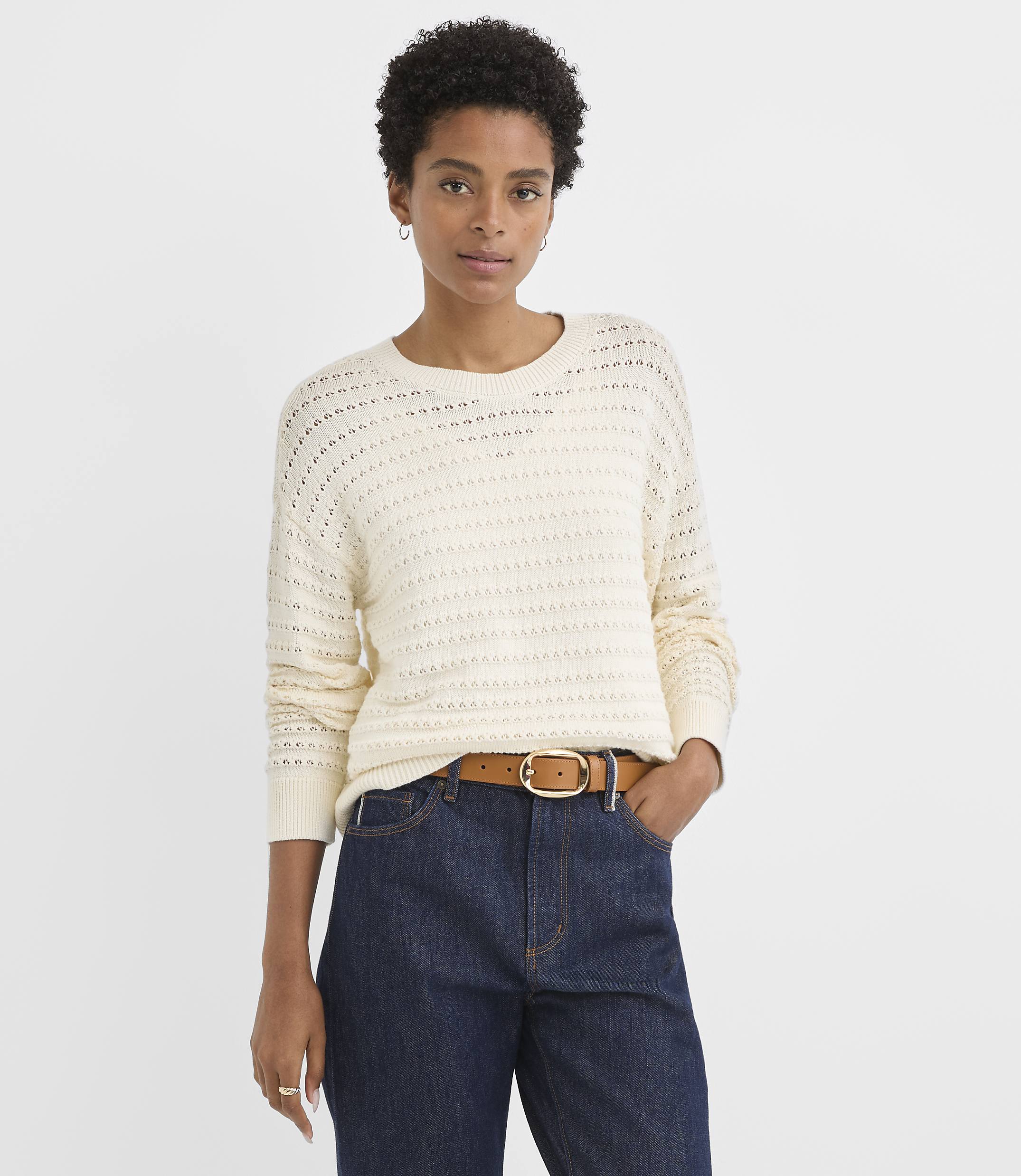 A woman in a cream-colored, open-knit sweater and dark wash jeans stands against a plain white background.