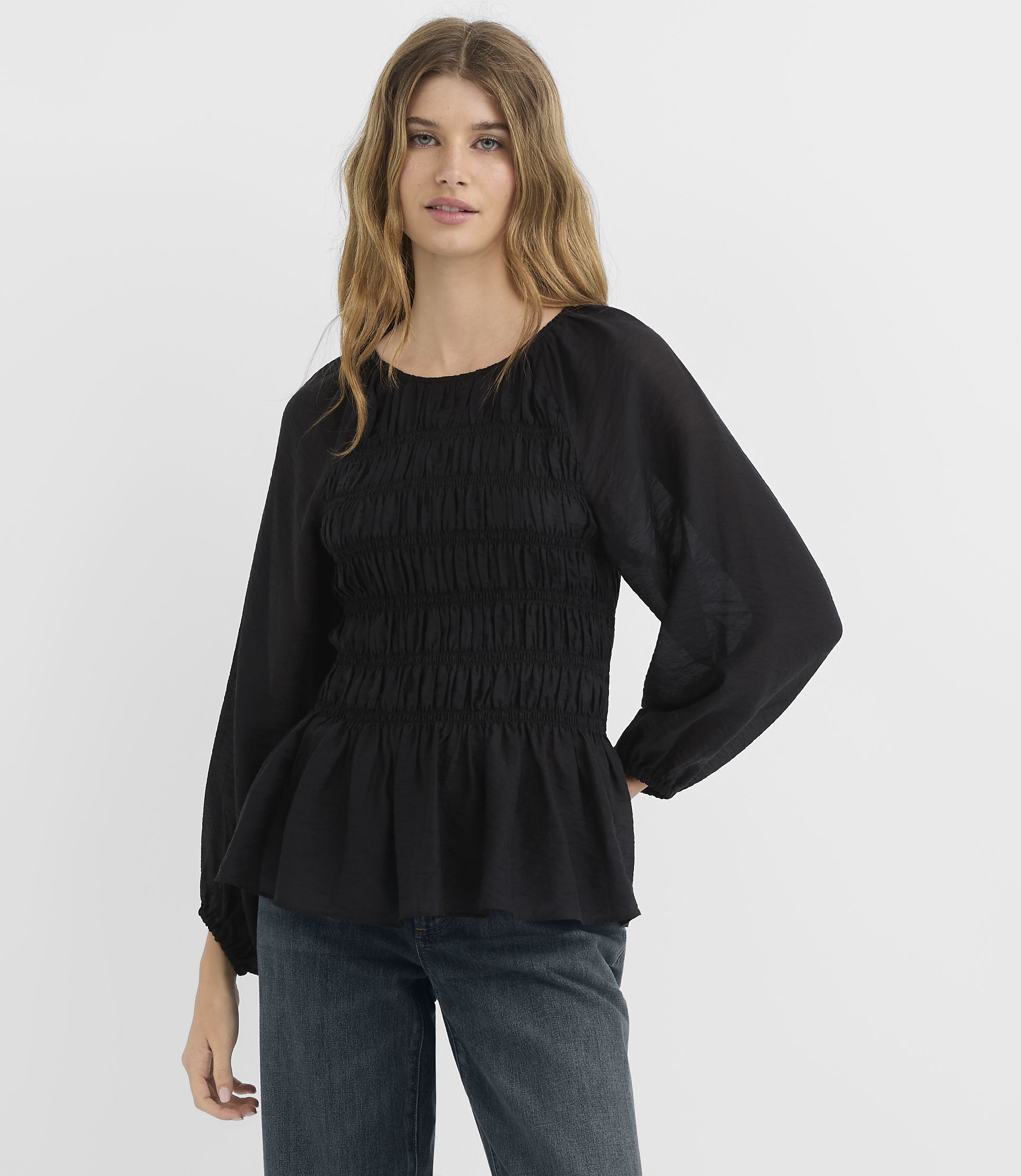A woman in a black smocked-bodice blouse and jeans stands against a plain white background.