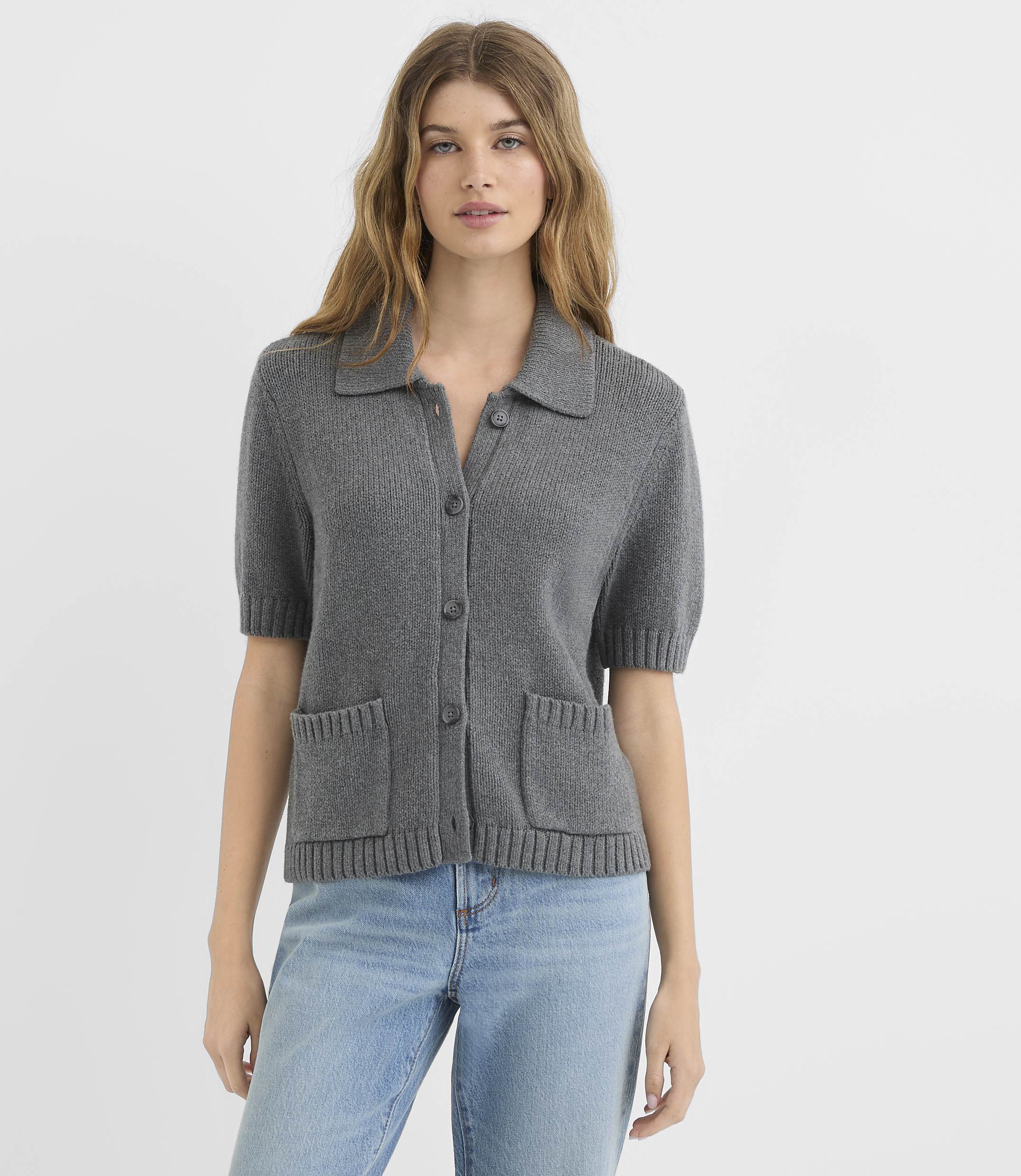 A woman in a gray short-sleeved collared cardigan and blue jeans stands against a plain white background.