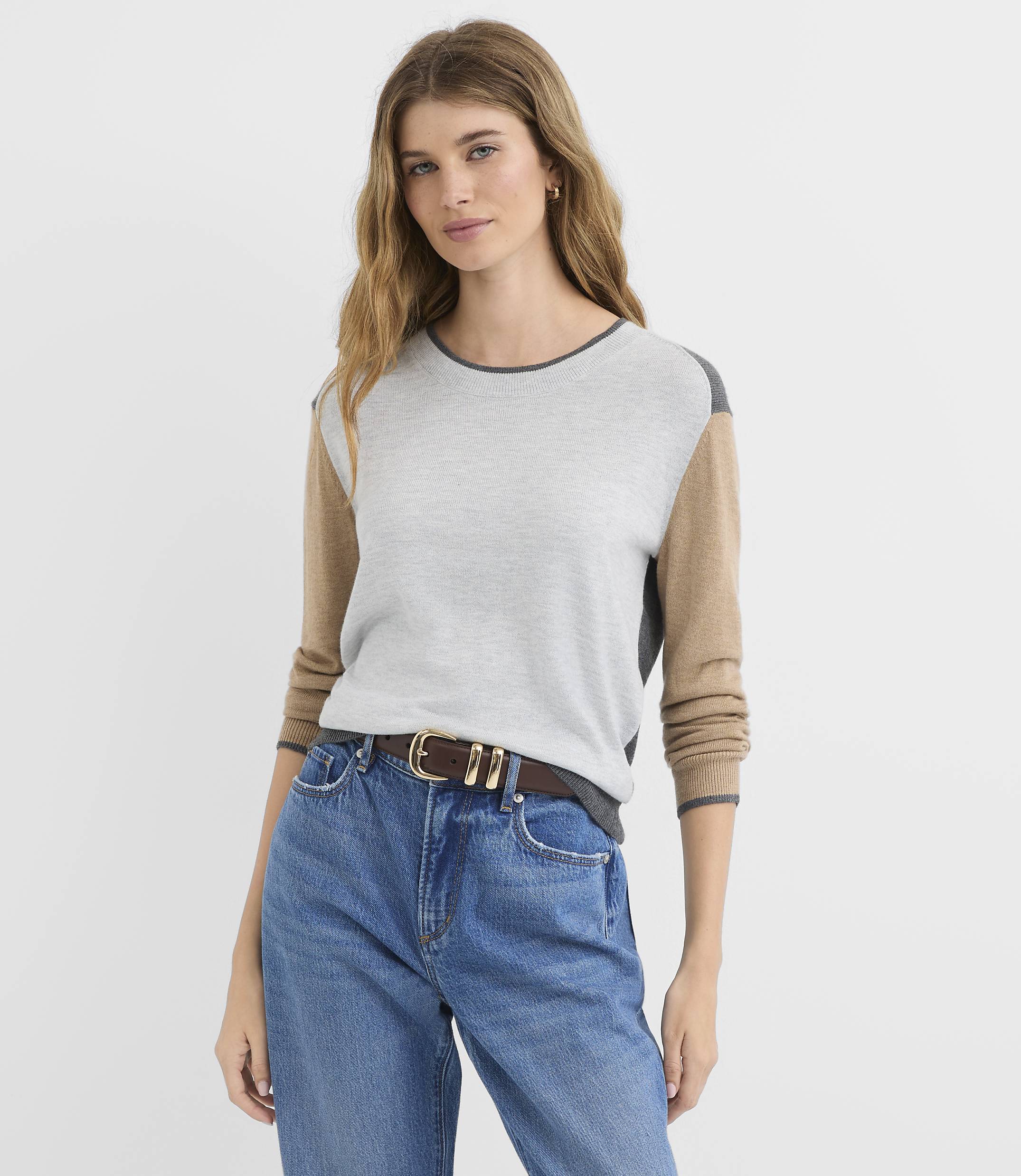 A woman in a color-blocked sweater and jeans stands against a plain gray background.