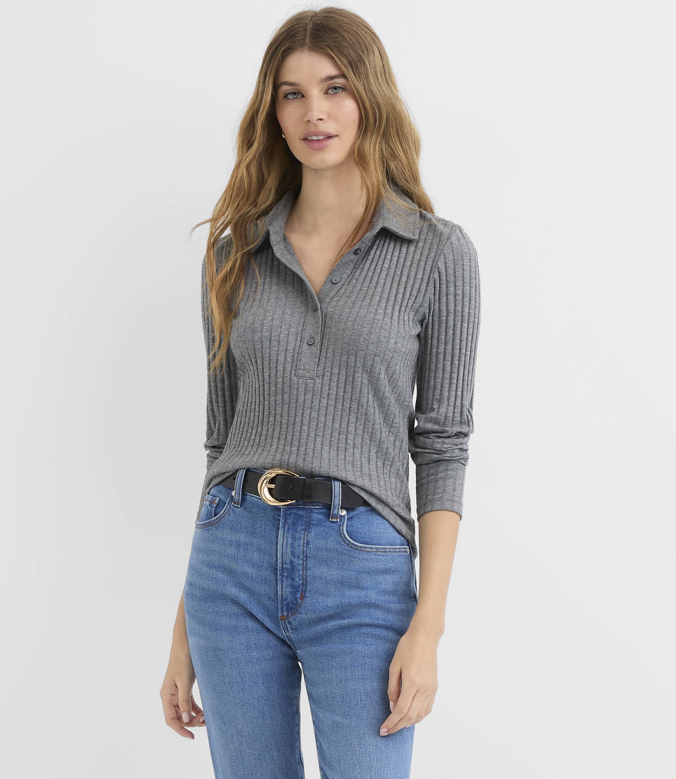 A woman in a grey ribbed polo shirt and blue jeans stands against a plain white background.