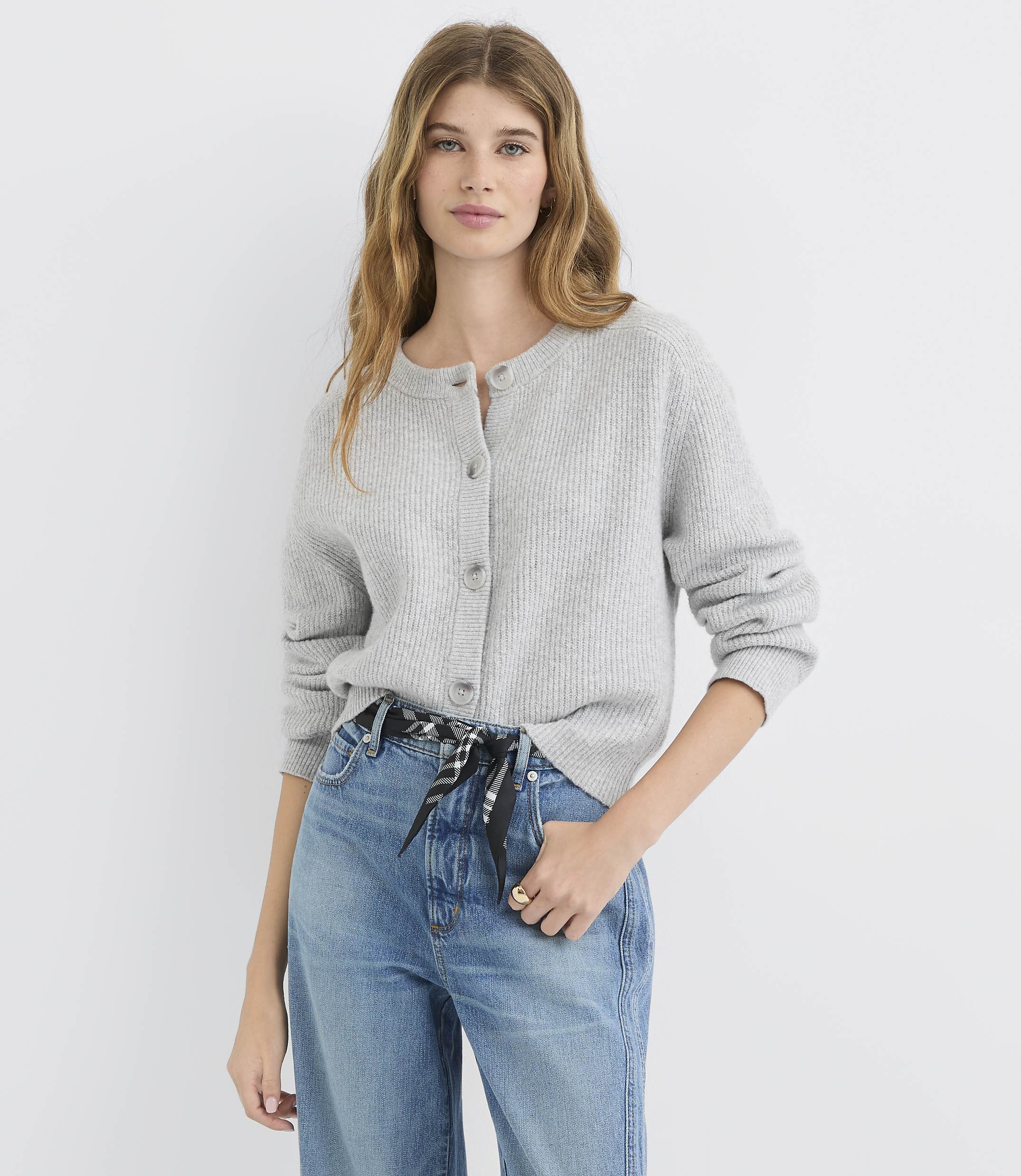 A woman in a grey cardigan and blue jeans stands against a plain white background.