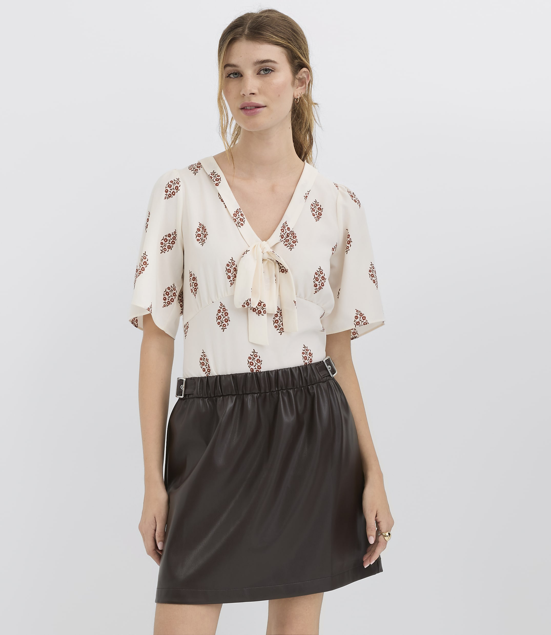 A woman in a patterned blouse and a dark skirt stands against a plain white background.
