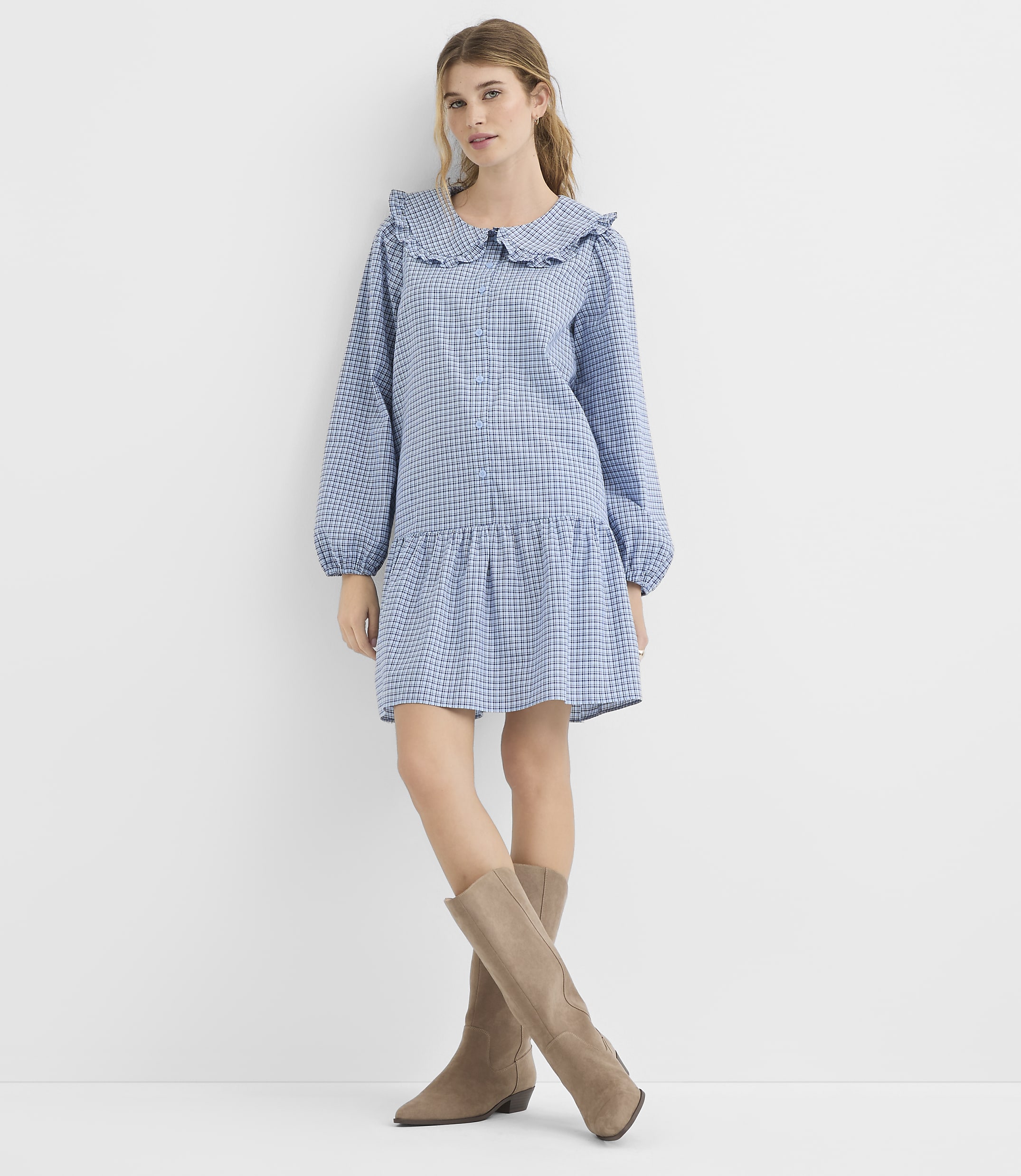 A young woman in a blue gingham dress and taupe boots stands against a plain white background.