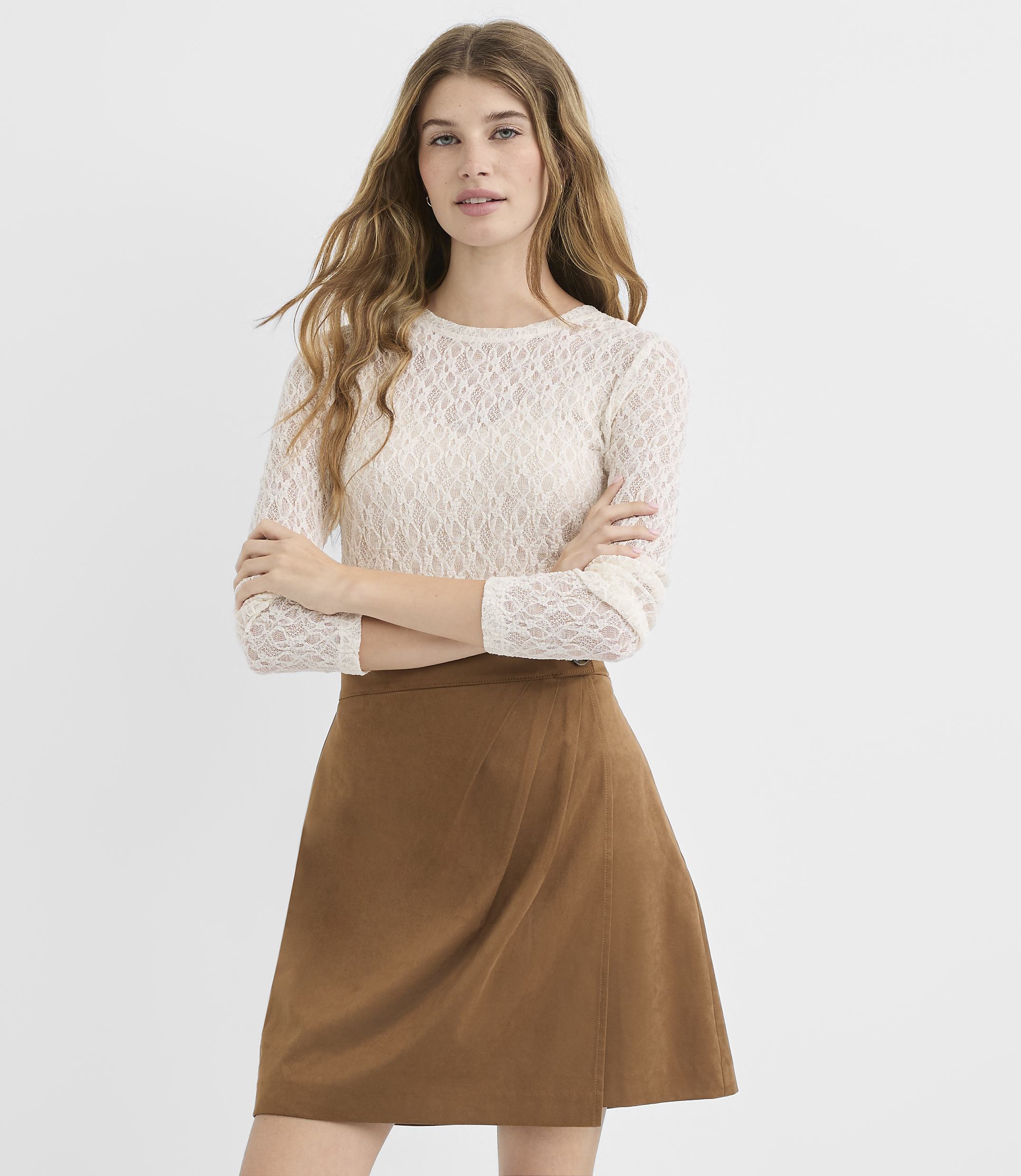 A woman in a white lace top and brown skirt stands with her arms crossed against a plain white background.