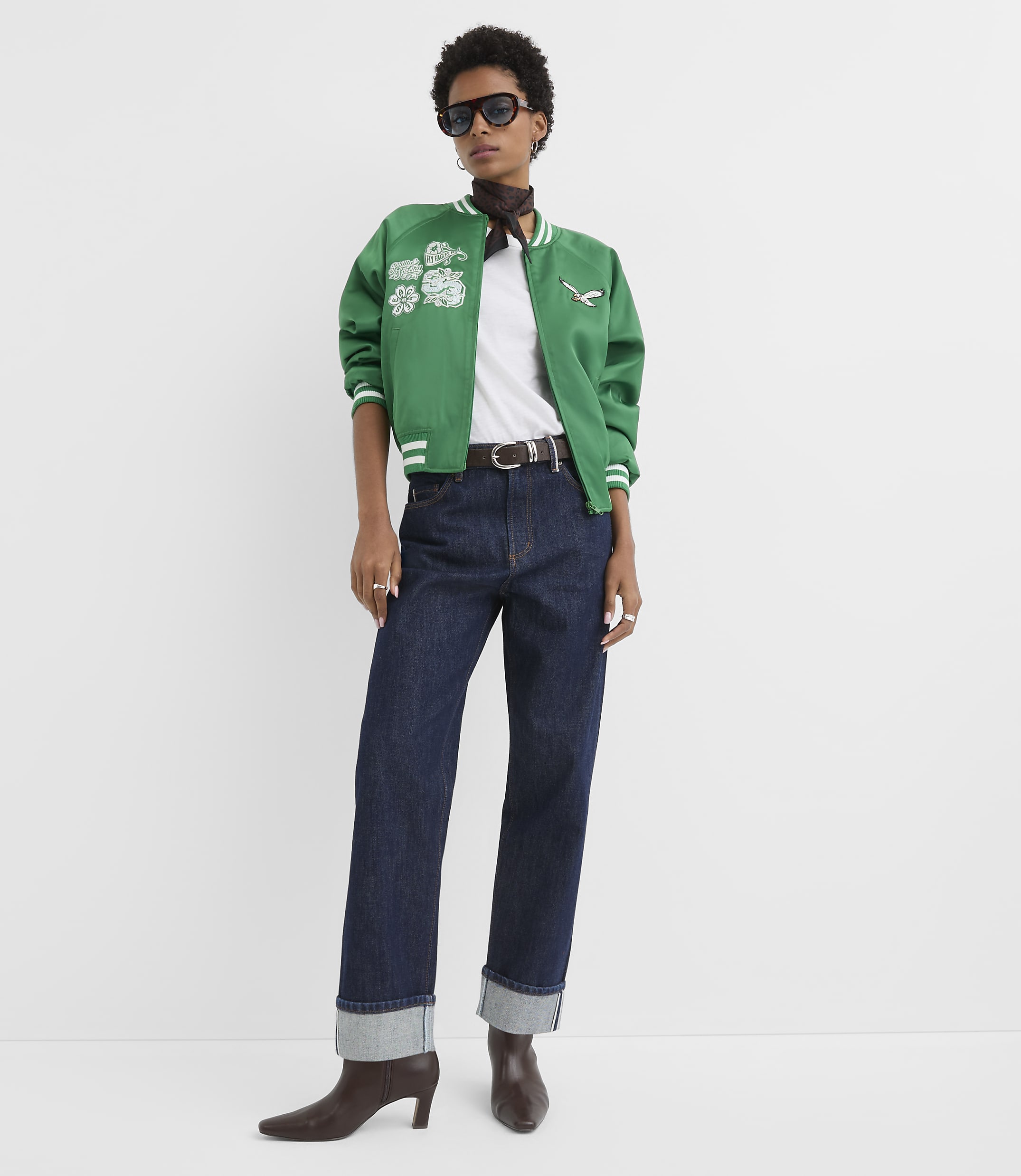 A woman in a green bomber jacket and dark jeans stands against a plain white background.
