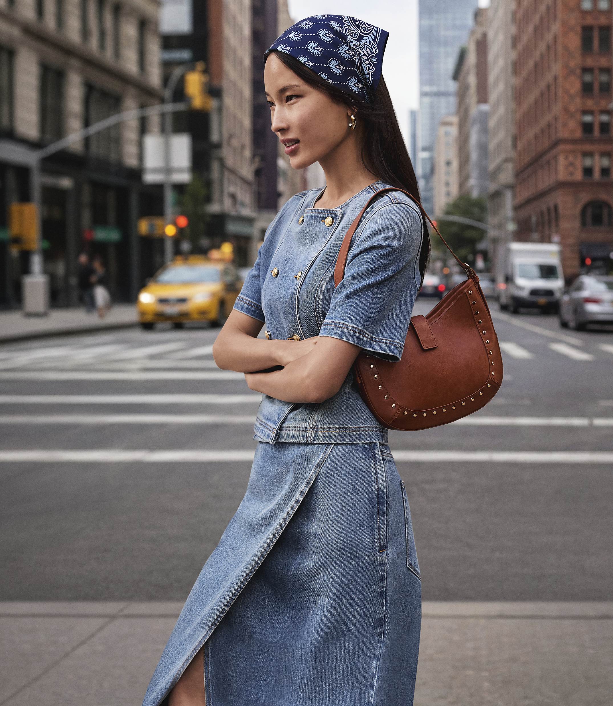 A woman in a denim outfit and bandana stands on a city street with a yellow taxi and buildings in the background.