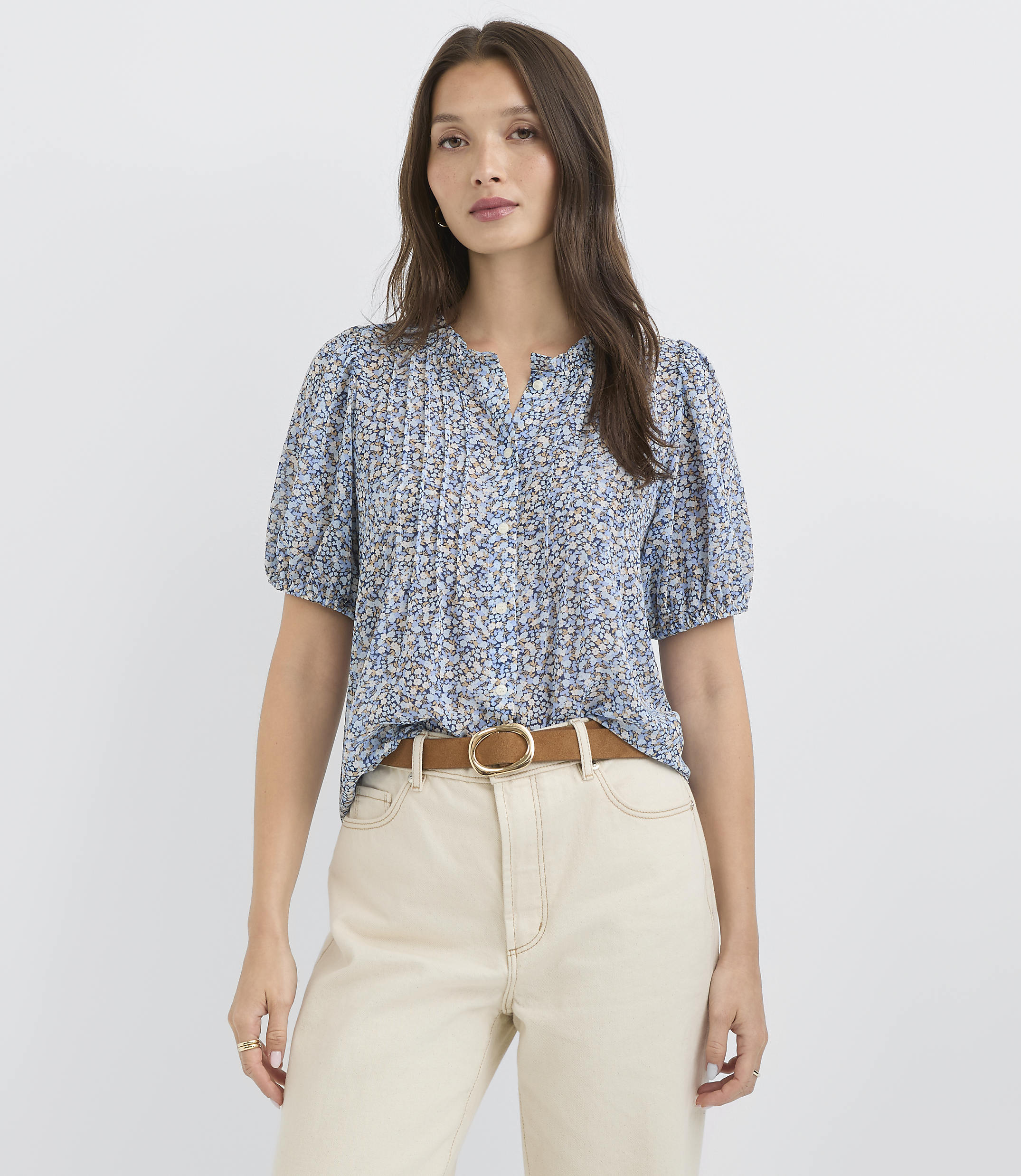 A woman in a floral blouse and cream pants stands against a plain white background.