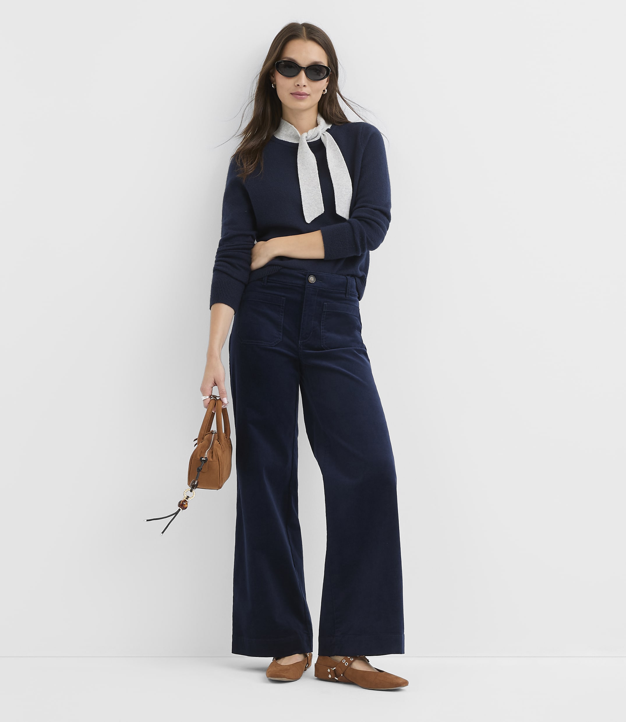 A woman in a navy sweater and wide-leg corduroy pants stands against a plain white background, holding a small brown purse.