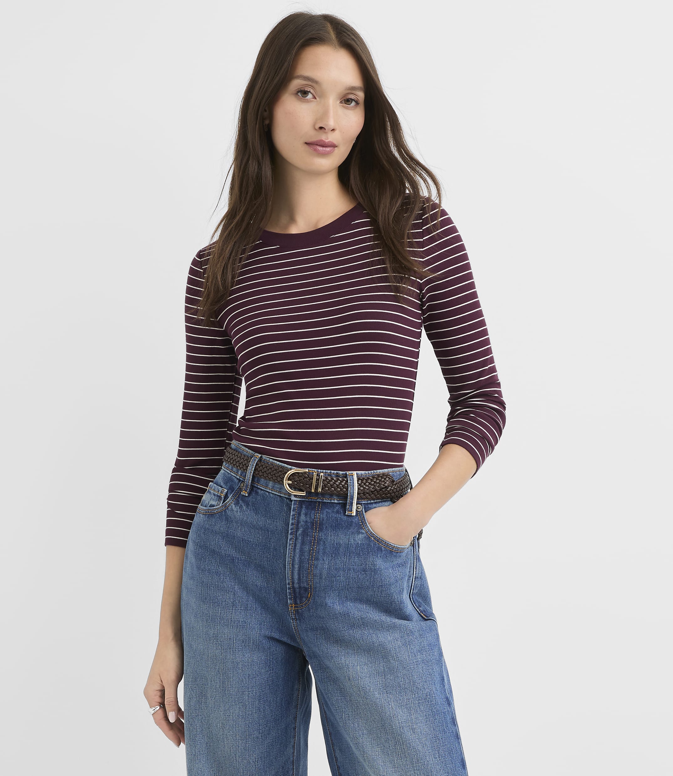 A woman in a striped shirt and jeans stands against a plain white background.