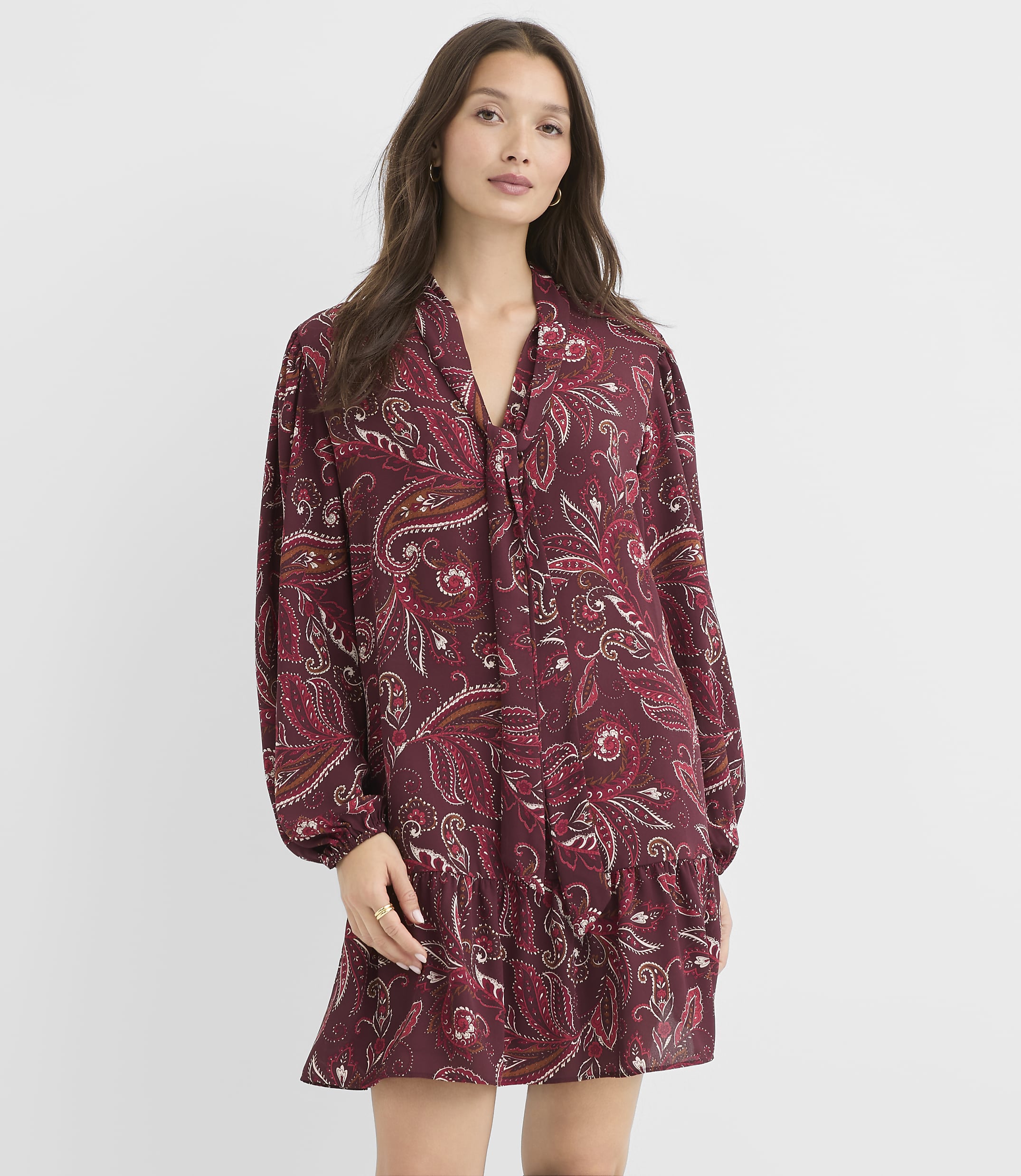 A woman in a burgundy paisley print dress stands against a plain white background.