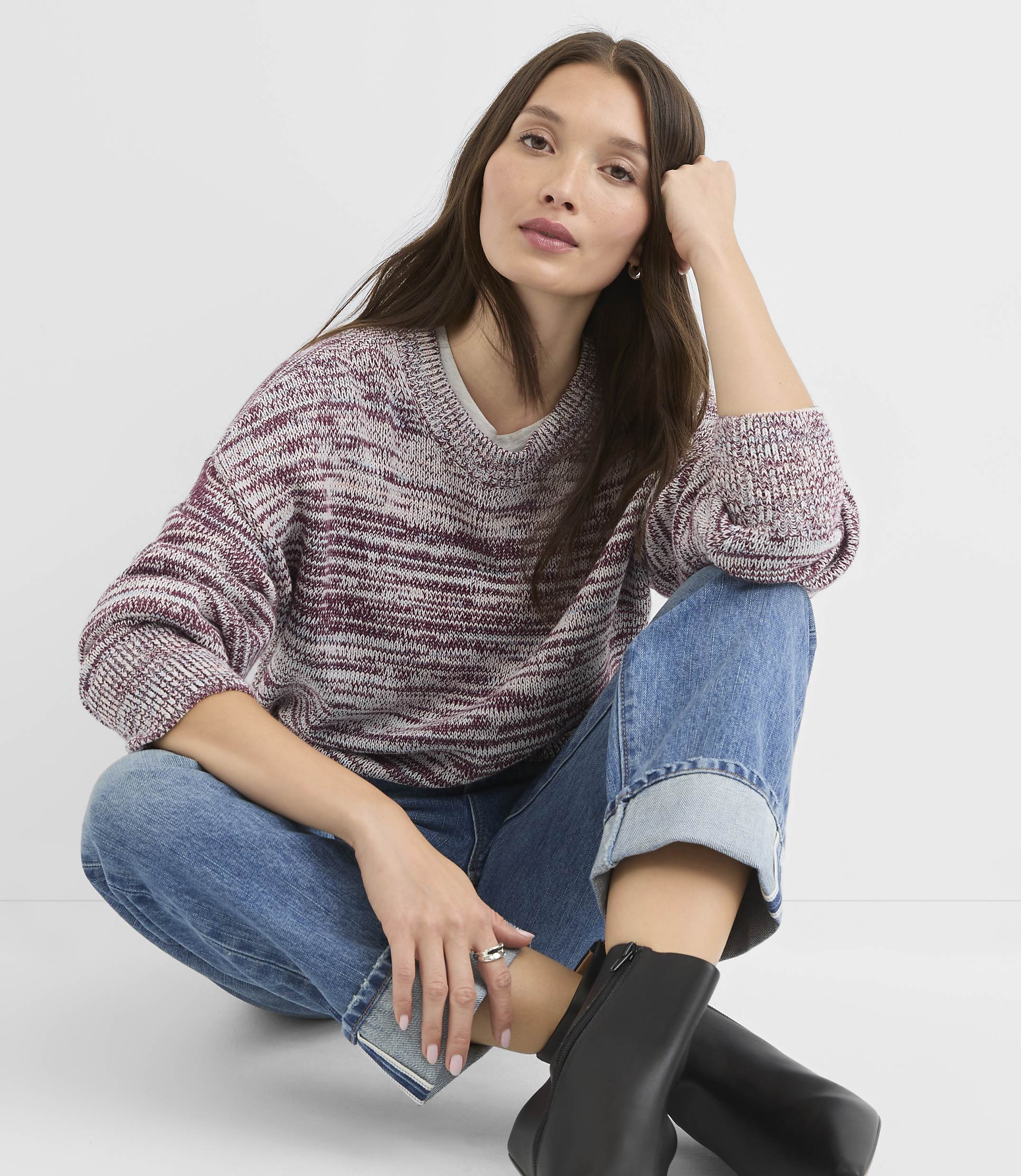 A woman in a marled burgundy and white sweater and cuffed blue jeans sits on a white background, leaning her head on her hand.