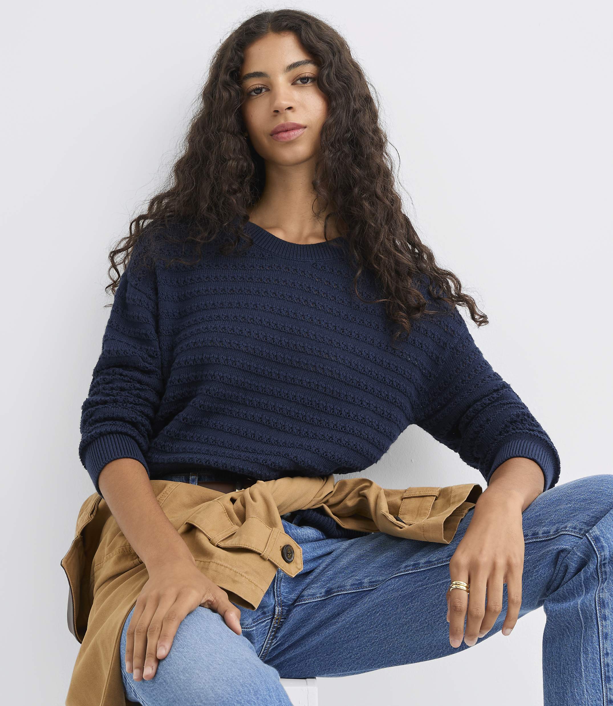 A woman with long, dark, curly hair wears a navy blue textured sweater and jeans, with a tan jacket draped over her shoulders, against a plain white background.