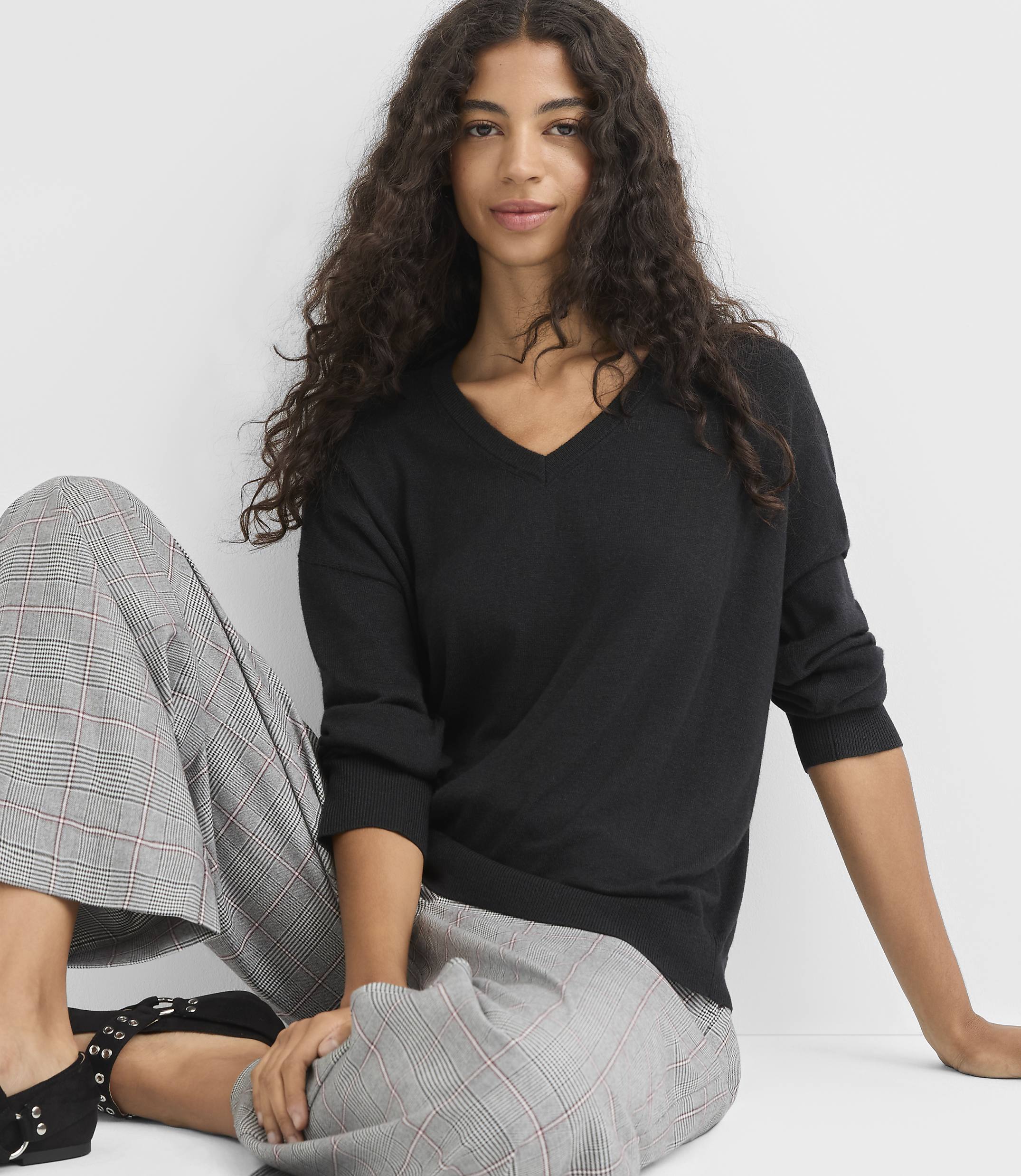 A woman with long, dark, curly hair sits in the foreground wearing a black v-neck sweater and plaid pants, with a plain white background behind her.