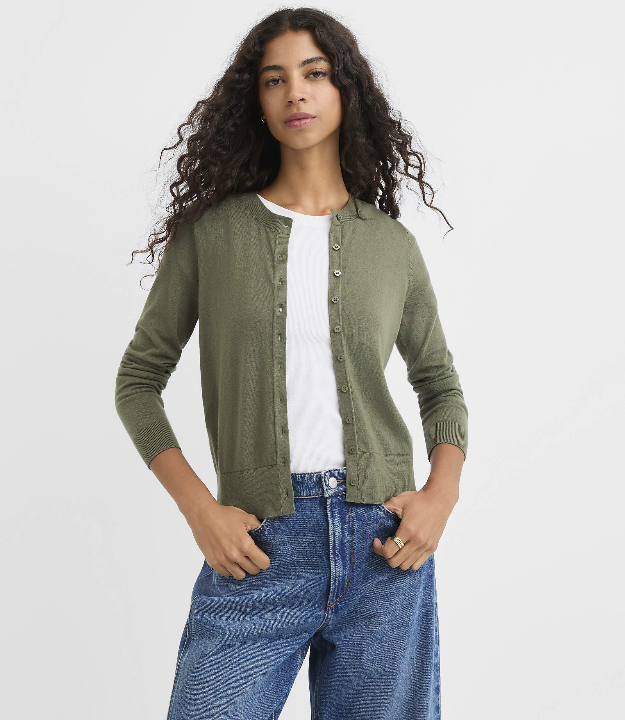 A woman in a green cardigan and blue jeans stands against a plain white background.