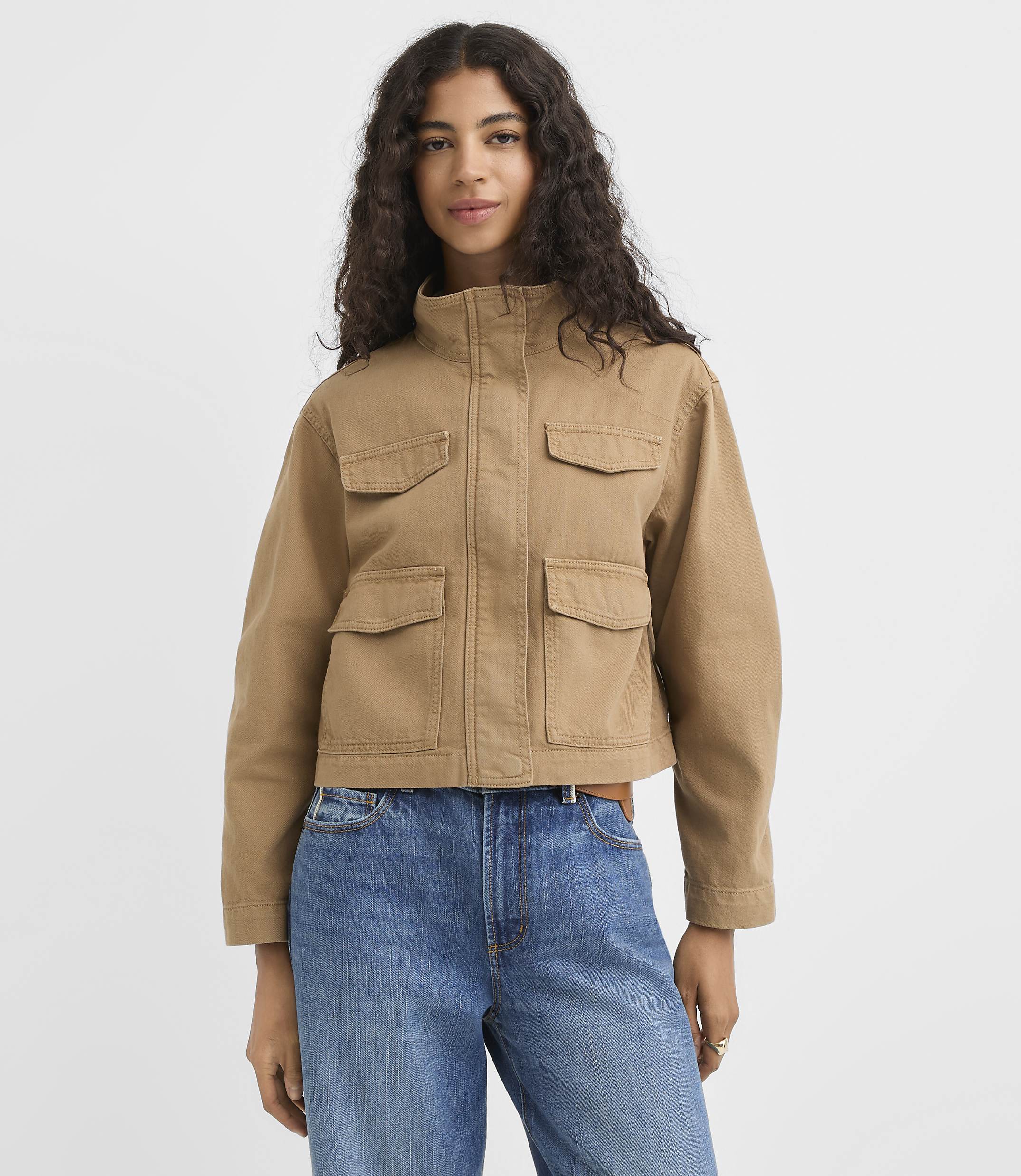 A young woman in a brown jacket and striped shirt stands against a plain white background.