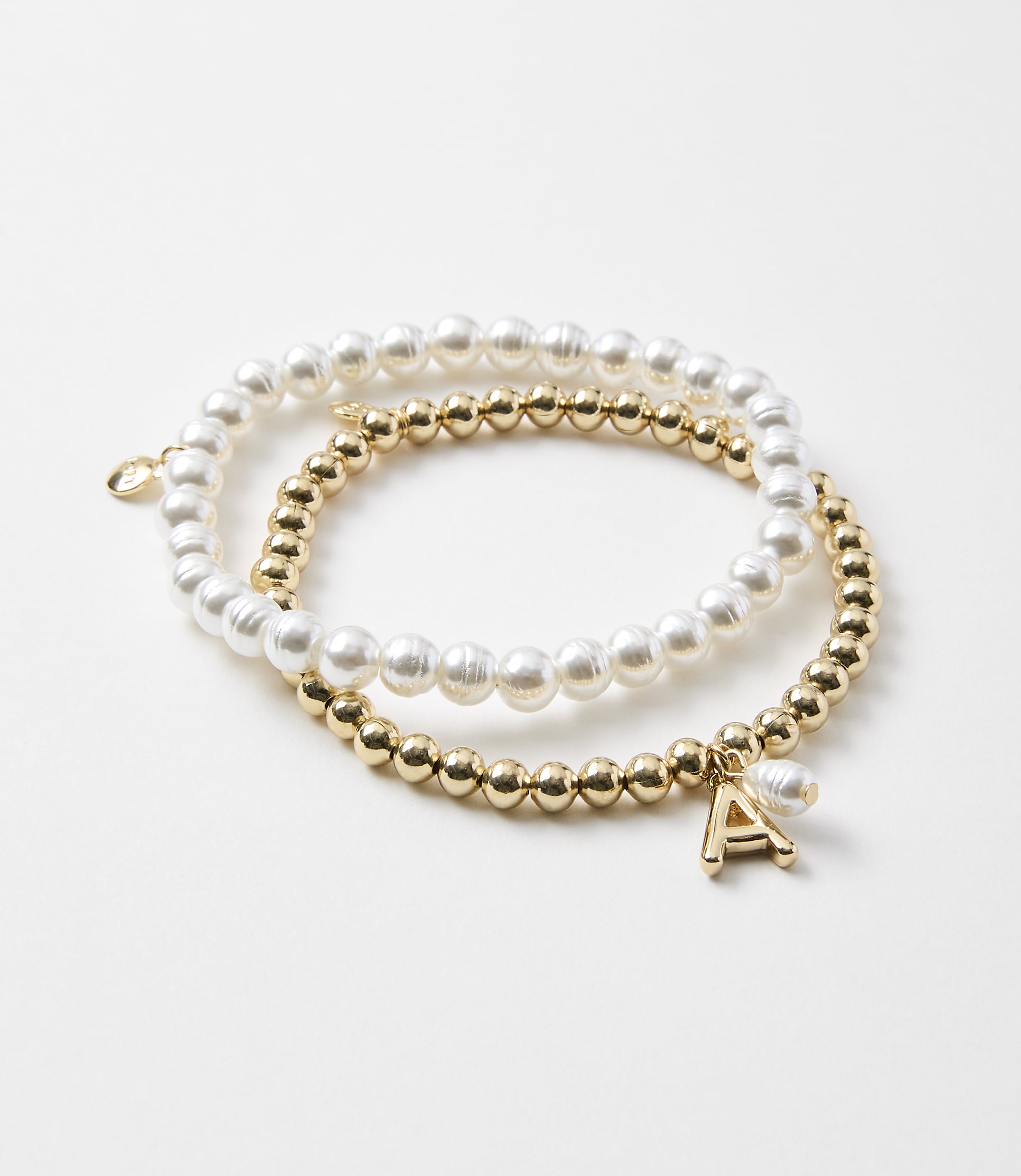 Two bracelets, one with pearls and the other with gold beads and an initial charm, rest on a plain white background.
