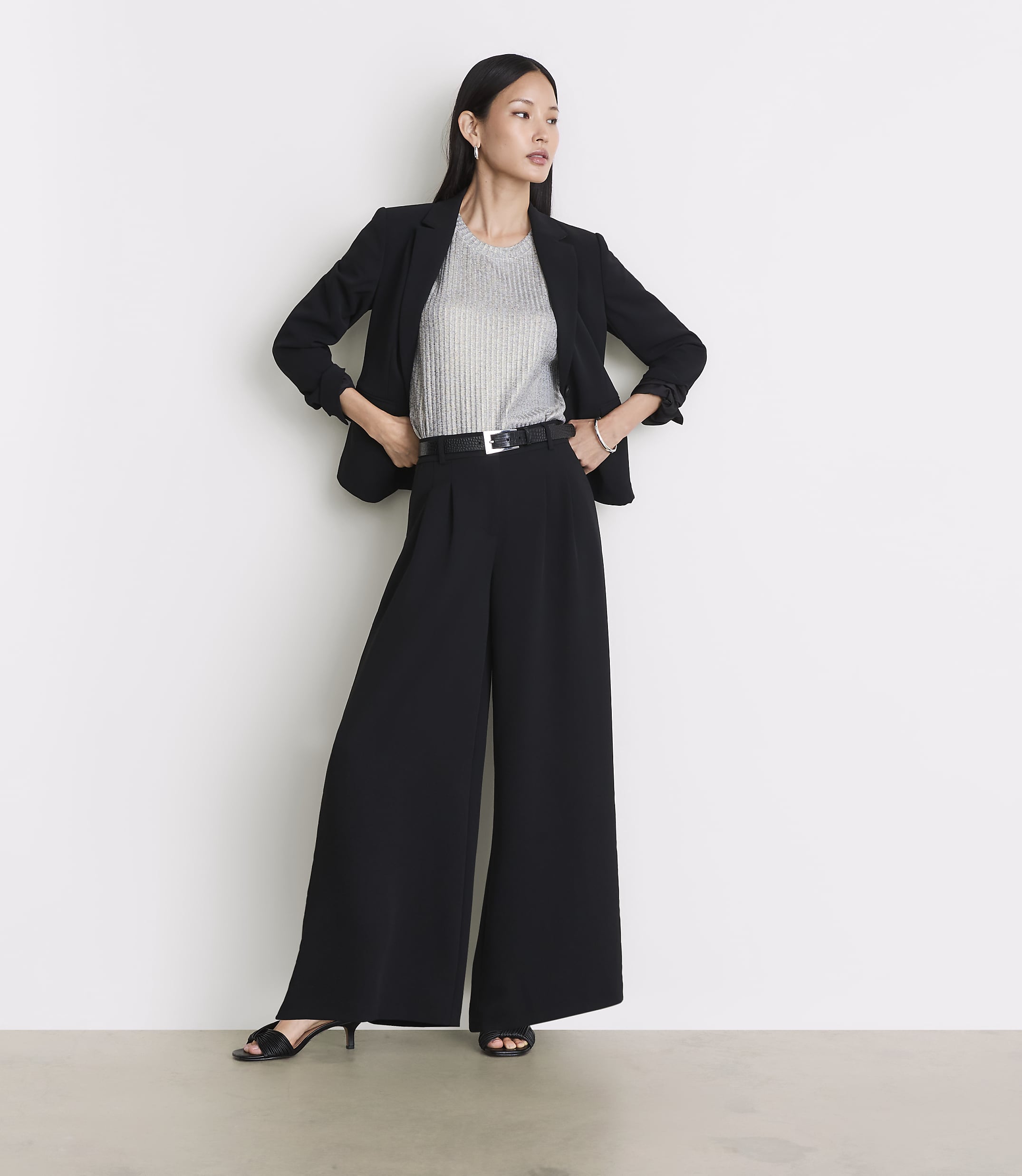 A woman in a black blazer and wide-leg pants stands against a plain white background.