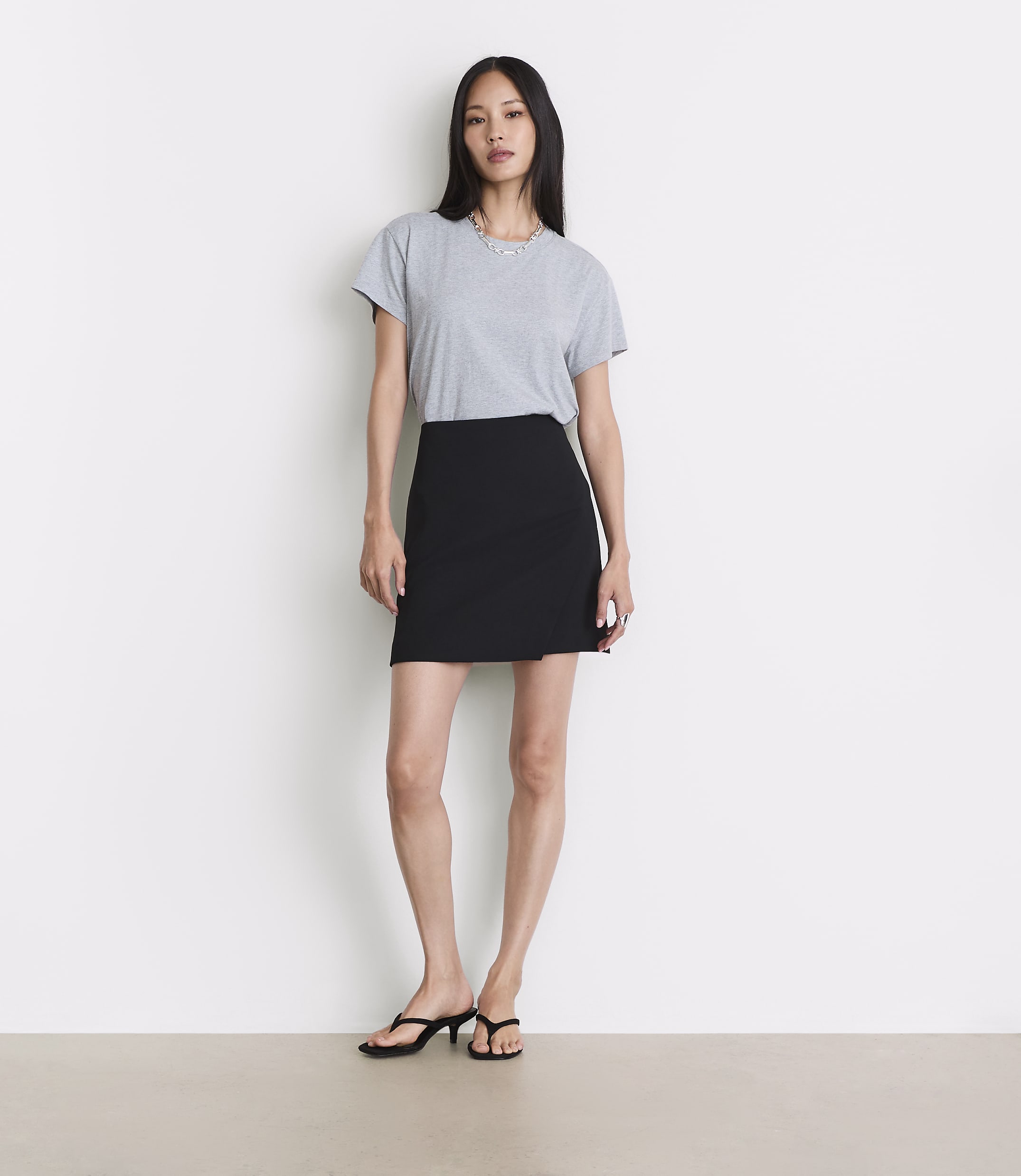 A woman in a grey t-shirt and black skirt stands against a plain white background.