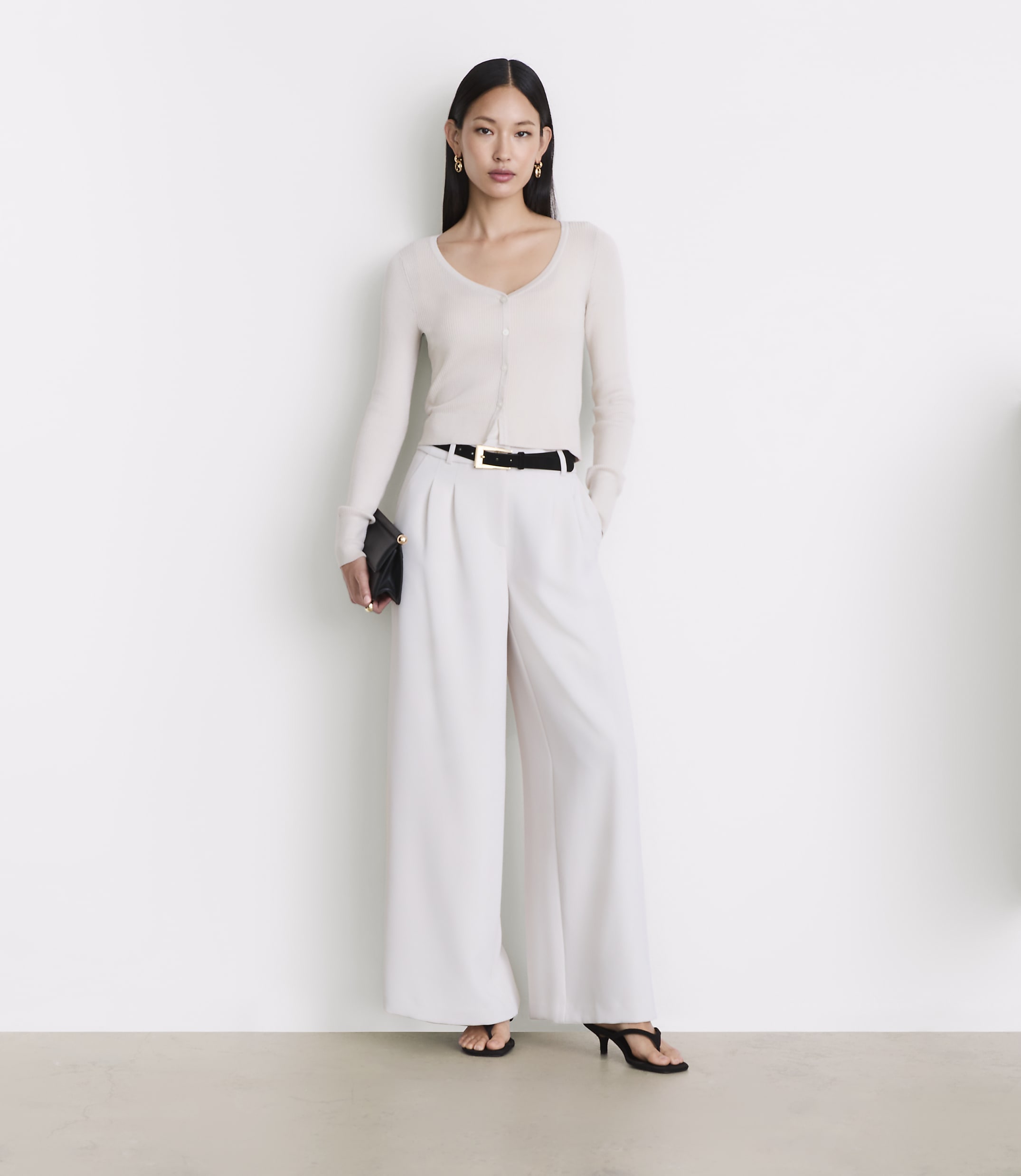 A woman in a cream cardigan and white wide-leg pants stands against a plain white background.