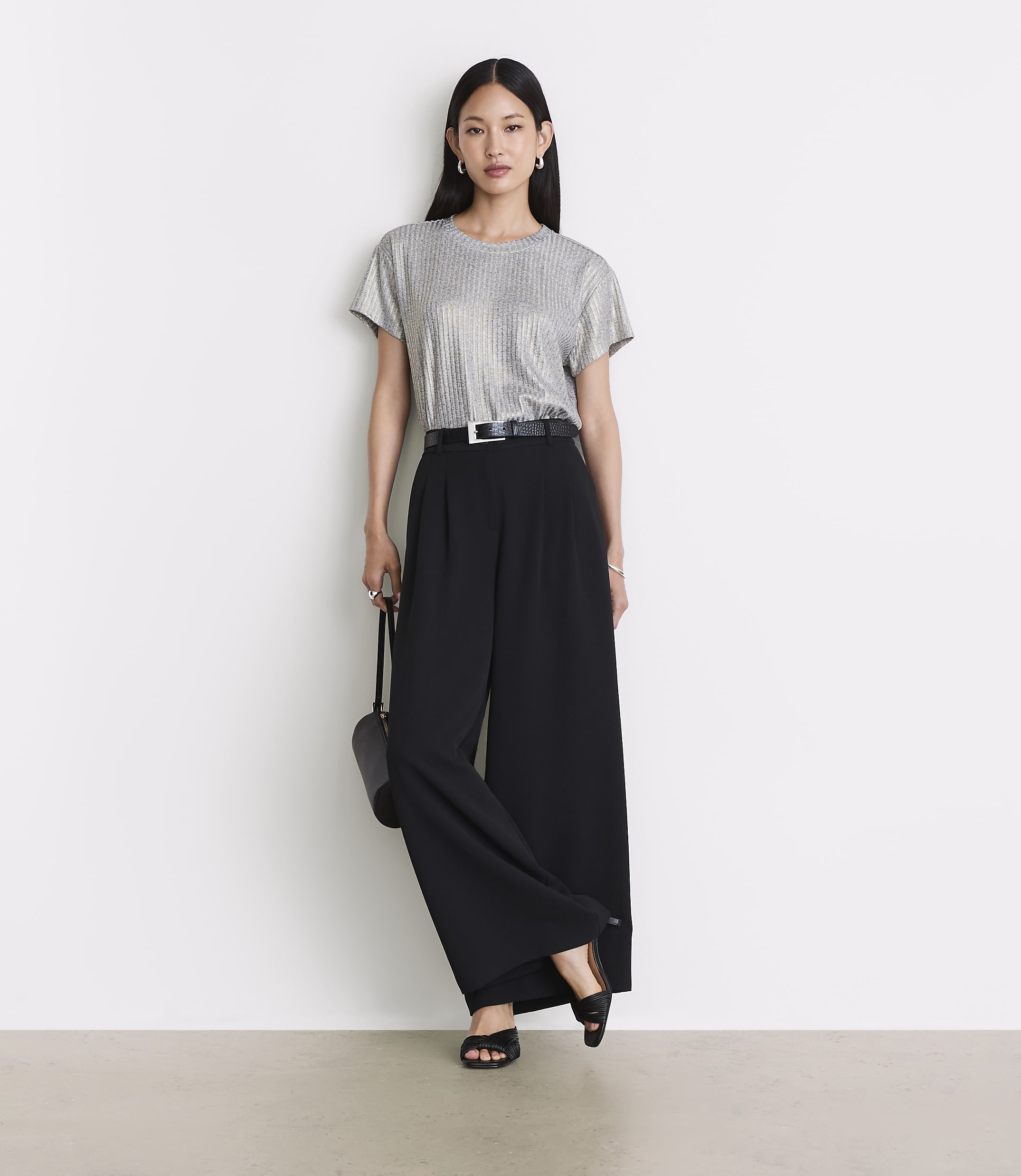 A woman in a sparkly silver top and wide-leg black pants stands against a plain white wall with a light-colored floor.