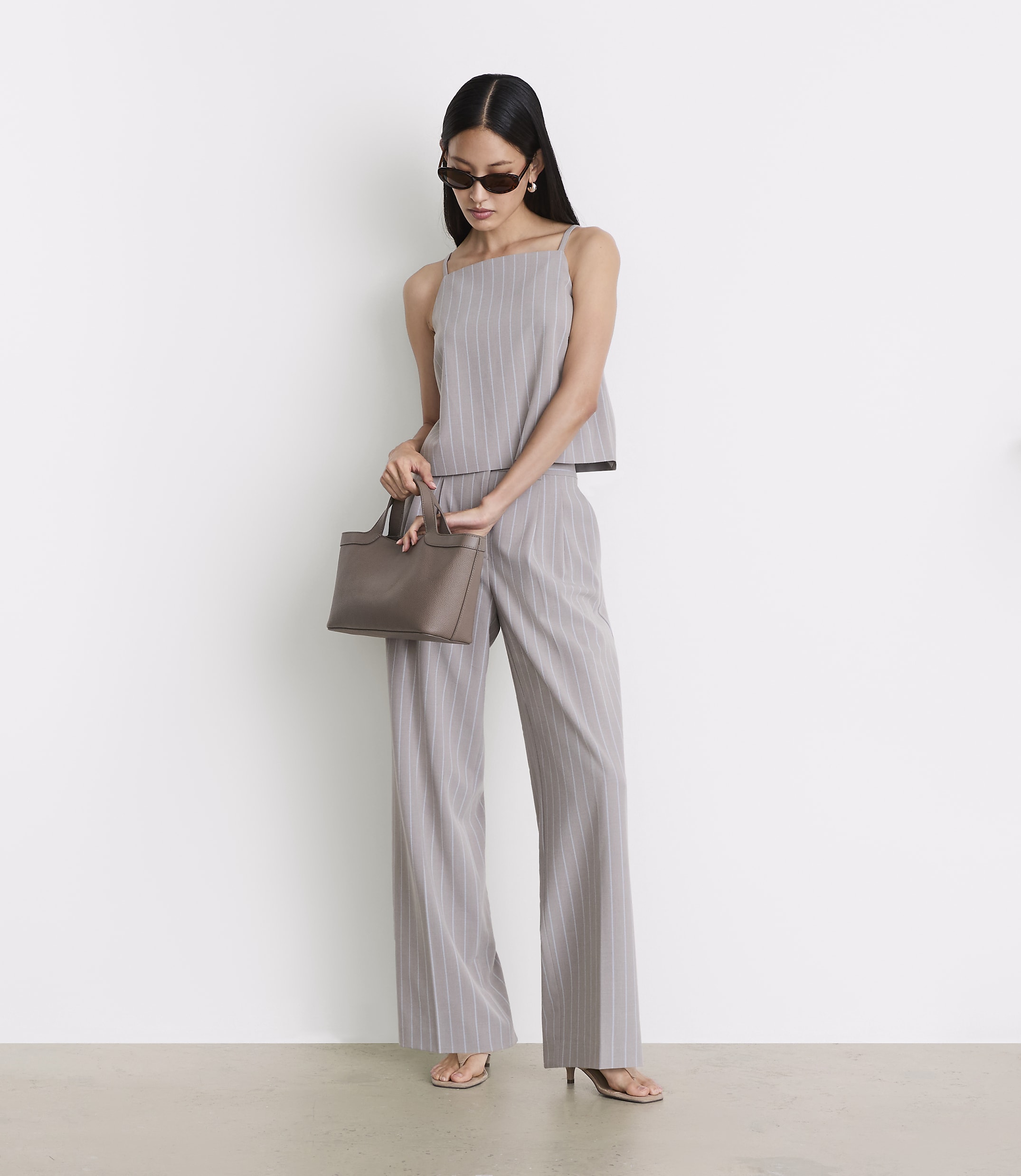 A woman in a pinstripe outfit and sunglasses holds a taupe handbag in front of a plain white background.