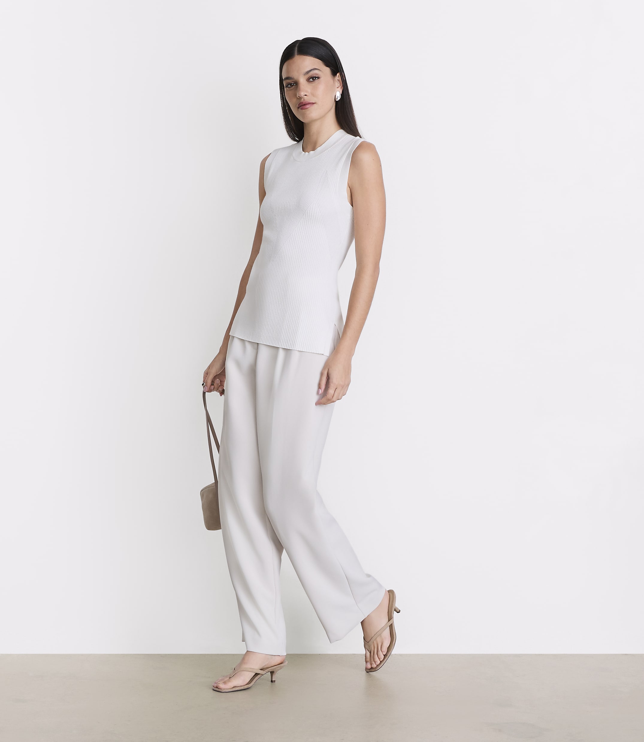 A woman in a white sleeveless top and wide-leg pants stands against a plain white background, holding a small beige purse.